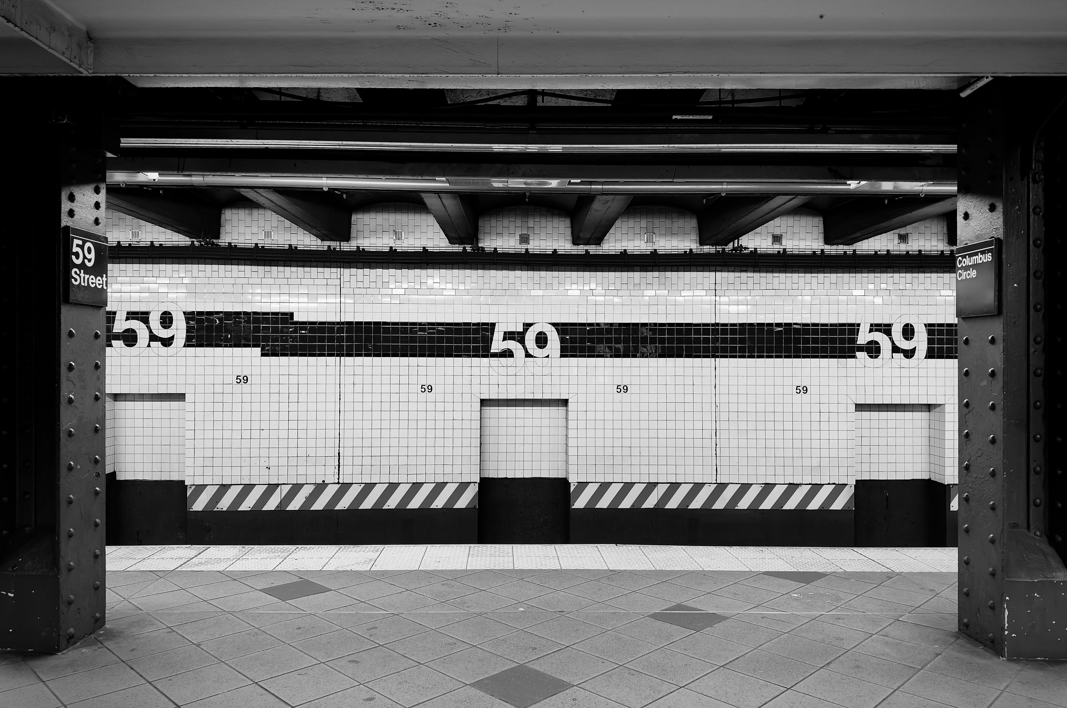 A noir, wide-angle photograph in landscape orientation that captures an empty subway platform at the 59th Street–Columbus Circle station in Manhattan. The shot is framed symmetrically between two dark, riveted steel girders in the foreground, creating a "window" effect that peers across the tracks toward the wall opposite the platform. The composition is defined by strong horizontal lines made up of the dark ceiling beams, and the white tiled walls intersected by a thick black horizontal band, and a repetitive pattern of diagonal hazard stripes along the platform's base. The lighting is crisp and moody, casting deep shadows in the recesses of the ceiling and under the girders, while the bright white tiles reflect a stark, artificial glow. The focal point of the image is the rhythmic repetition of the number "59" across the station wall. Large, bold white numerals are embedded within the black tile band, while smaller versions of the number are printed on the white tiles below them. To the left, a vertical black sign with white text reads "59 Street," and to the right, a matching sign reads "Columbus Circle." The ground in the foreground shows a diamond-patterned tile floor, and the entire scene is devoid of people, heightening a sense of urban solitude and noir-inspired stillness.