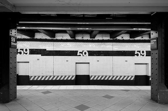 A noir, wide-angle photograph in landscape orientation that captures an empty subway platform at the 59th Street–Columbus Circle station in Manhattan. The shot is framed symmetrically between two dark, riveted steel girders in the foreground, creating a "window" effect that peers across the tracks toward the wall opposite the platform. The composition is defined by strong horizontal lines made up of the dark ceiling beams, and the white tiled walls intersected by a thick black horizontal band, and a repetitive pattern of diagonal hazard stripes along the platform's base. The lighting is crisp and moody, casting deep shadows in the recesses of the ceiling and under the girders, while the bright white tiles reflect a stark, artificial glow. The focal point of the image is the rhythmic repetition of the number "59" across the station wall. Large, bold white numerals are embedded within the black tile band, while smaller versions of the number are printed on the white tiles below them. To the left, a vertical black sign with white text reads "59 Street," and to the right, a matching sign reads "Columbus Circle." The ground in the foreground shows a diamond-patterned tile floor, and the entire scene is devoid of people, heightening a sense of urban solitude and noir-inspired stillness.