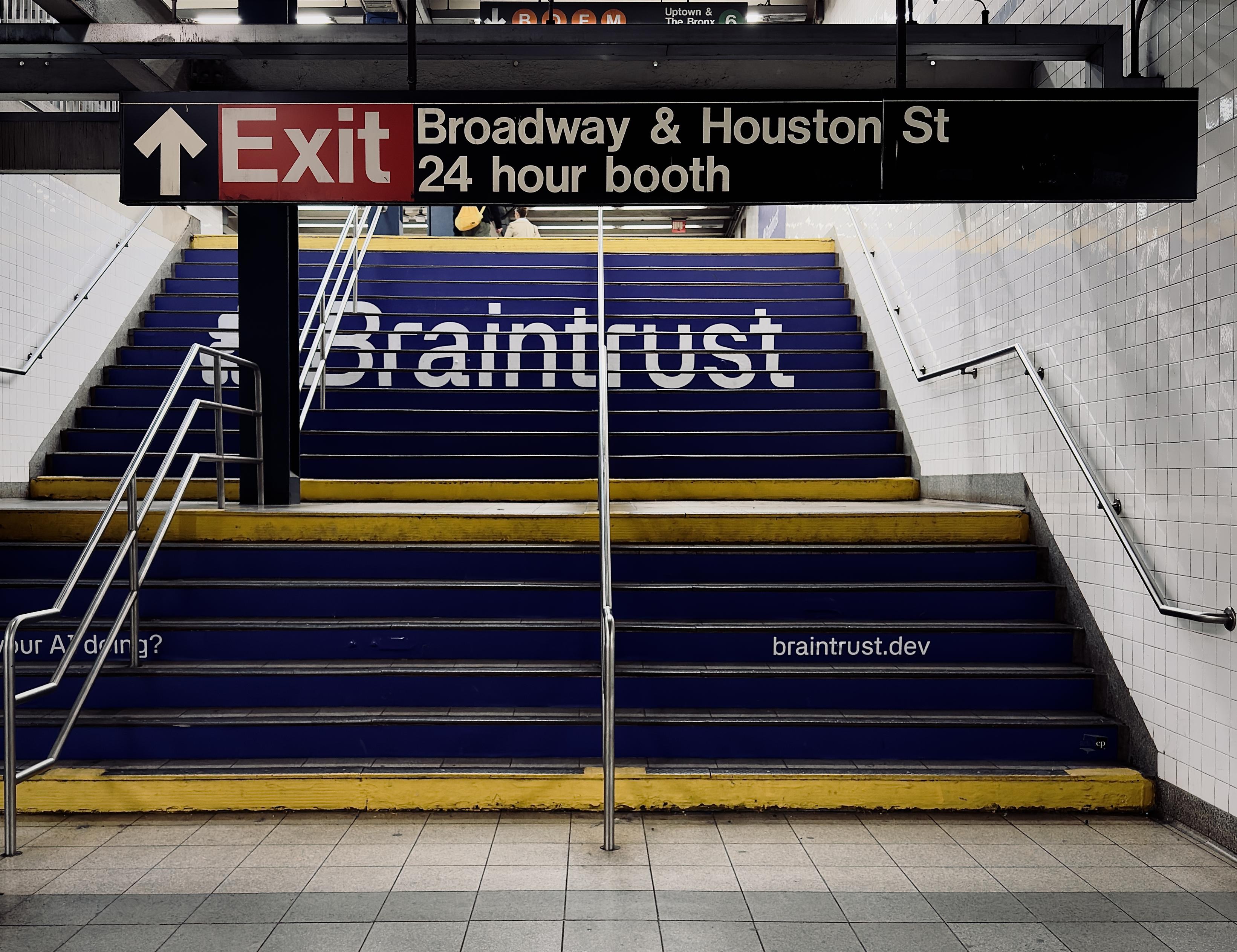 A color, wide-angle photograph in 5:4 orientation of the Broadway–Lafayette subway station in Manhattan. The focal point of the scene is a wide set of stairs leading toward an exit. The staircase is transformed by an advertisement for Braintrust, with the risers of the steps painted a deep navy blue. Spanned across the upper half of the stairs in large, bold white sans-serif letters is the word "Braintrust." Below this, on a lower section of the steps, the URL "braintrust.dev" is printed in smaller white text on the right side, the partial phrase "your AI doing?" is visible on the left. The top and bottom steps of each flight are accented with a bright yellow safety strip, creating a sharp contrast against the dark blue advertisement and the light grey tiled floor in the foreground. Above the stairs, a black rectangular directional sign hangs from the ceiling, featuring white text that reads "Exit" in a large font next to an upward-pointing arrow. To the right of the exit text, the sign indicates "Broadway & Houston St" and "24 hour booth." Above this main sign, further up the stairs is a smaller sign with colorful circular icons for the B, D, F, and M subway lines visible, along with directions for "Uptown & The Bronx" and the green 6 line. The station walls are covered in classic white rectangular subway tiles, and several silver metal handrails run up the center and sides of the stairs. The lighting is even and industrial.