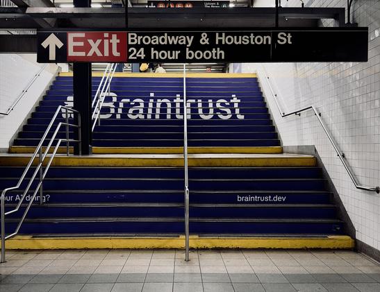 A color, wide-angle photograph in 5:4 orientation of the Broadway–Lafayette subway station in Manhattan. The focal point of the scene is a wide set of stairs leading toward an exit. The staircase is transformed by an advertisement for Braintrust, with the risers of the steps painted a deep navy blue. Spanned across the upper half of the stairs in large, bold white sans-serif letters is the word "Braintrust." Below this, on a lower section of the steps, the URL "braintrust.dev" is printed in smaller white text on the right side, the partial phrase "your AI doing?" is visible on the left. The top and bottom steps of each flight are accented with a bright yellow safety strip, creating a sharp contrast against the dark blue advertisement and the light grey tiled floor in the foreground. Above the stairs, a black rectangular directional sign hangs from the ceiling, featuring white text that reads "Exit" in a large font next to an upward-pointing arrow. To the right of the exit text, the sign indicates "Broadway & Houston St" and "24 hour booth." Above this main sign, further up the stairs is a smaller sign with colorful circular icons for the B, D, F, and M subway lines visible, along with directions for "Uptown & The Bronx" and the green 6 line. The station walls are covered in classic white rectangular subway tiles, and several silver metal handrails run up the center and sides of the stairs. The lighting is even and industrial.