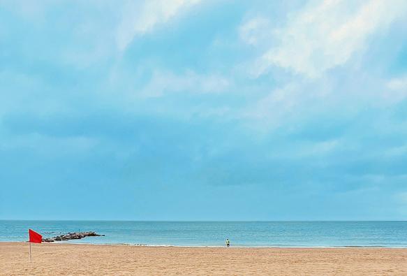 A color, wide-angle photograph that captures an expansive, minimalist view of a beach at Coney Island on a gloomy, overcast day. In the foreground, a vast expanse of tan sand stretches across the bottom third of the frame, showing faint patterns of footprints and tractor treads. A single bright red flag on a thin pole stands prominently on the left side of the sand, providing a sharp contrast to the muted surroundings. In the mid-ground, a small rock jetty extends from the left into the calm, pale blue water. Further along the shoreline, a lone figure wearing a yellow safety vest stands at the water's edge, appearing very small against the scale of the environment. The upper two-thirds of the photograph are dominated by a massive, hazy sky filled with thick, soft blue and grey clouds. The horizon line is flat and sits low in the frame, creating a sense of isolation and immense open space. The overall lighting is soft and diffused, lacking harsh shadows, which gives the scene a quiet, desolate, and almost painterly quality. The color palette is restricted to shades of sandy beige, soft cerulean, and the singular pop of red from the warning flag.