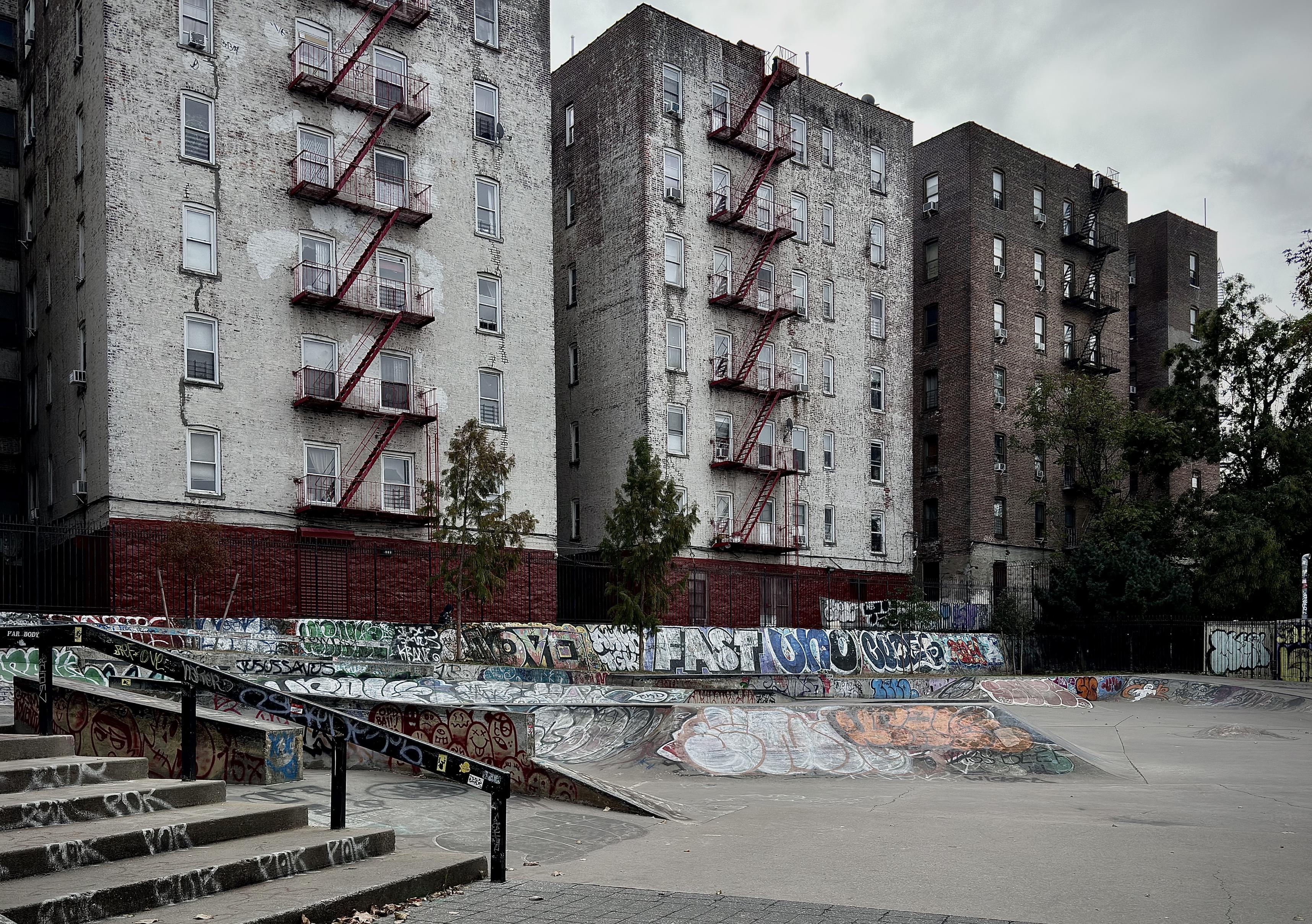A color, wide-angle photograph that depicts a gritty, urban skate park along River Avenue in the Highbridge neighborhood of the Bronx. It is an overcast morning, and the scene is characterized by extensive graffiti and its proximity to towering residential buildings. In the foreground, a set of concrete stairs with a dark metal handrail leads down to the skating area. The handrail and the side of the stairs are covered in various tags and stickers. The park itself features a smooth concrete surface with a large, curved bowl element, which is heavily layered with colorful, overlapping graffiti murals, tags, and street art in shades of blue, red, white, and orange. Behind the skate park, three large, multi-story brick apartment buildings rise against a grey, overcast sky. The two buildings on the left are painted white, though the paint is weathered and peeling, revealing the darker brick underneath. These buildings feature distinctive red metal fire escapes zigzagging down their facades. The third building to the right remains a traditional dark brown brick. Small, sparse green trees stand between the park and the buildings, partially obscured by a dark metal fence. The overall atmosphere is somber and cool.