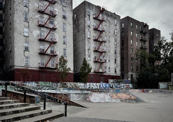 A color, wide-angle photograph that depicts a gritty, urban skate park along River Avenue in the Highbridge neighborhood of the Bronx. It is an overcast morning, and the scene is characterized by extensive graffiti and its proximity to towering residential buildings. In the foreground, a set of concrete stairs with a dark metal handrail leads down to the skating area. The handrail and the side of the stairs are covered in various tags and stickers. The park itself features a smooth concrete surface with a large, curved bowl element, which is heavily layered with colorful, overlapping graffiti murals, tags, and street art in shades of blue, red, white, and orange. Behind the skate park, three large, multi-story brick apartment buildings rise against a grey, overcast sky. The two buildings on the left are painted white, though the paint is weathered and peeling, revealing the darker brick underneath. These buildings feature distinctive red metal fire escapes zigzagging down their facades. The third building to the right remains a traditional dark brown brick. Small, sparse green trees stand between the park and the buildings, partially obscured by a dark metal fence. The overall atmosphere is somber and cool.