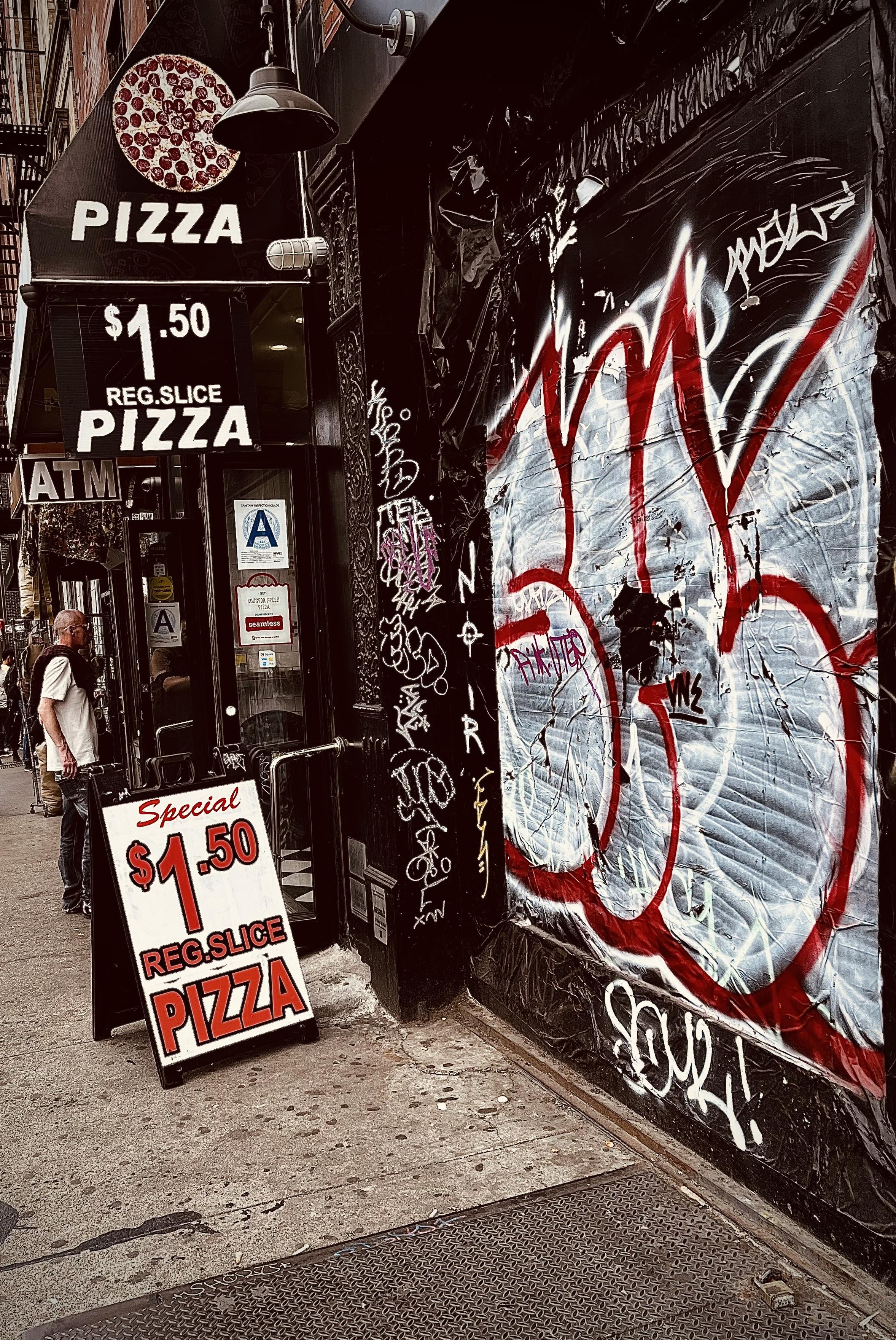 A color, wide-angle photograph in portrait orientation that captures a gritty, high-contrast street scene of a budget-friendly pizza shop on the Lower East Side of Manhattan. The shop's entrance is marked by multiple black signs and a sidewalk A-frame board advertising a "Special $1.50 Reg. Slice Pizza." Above the door, a black awning features a graphic of a pepperoni pizza and more bold white lettering. The storefront is narrow, with a visible "Grade A" health department rating in the window and an ATM sign protruding from the side. A man in a white t-shirt and glasses stands near the entrance, looking off-camera, while the sidewalk shows the typical wear and texture of a busy urban environment. The right side of the frame is dominated by a dark, textured wall heavily covered in street art. A large, stylized graffiti tag in bold red and white outlines sprawls across a corrugated metal shutter, layered over various smaller white and purple scrawls. The word "NOIR" is written vertically in white paint on a black pillar. The lighting is moody and dramatic, with deep shadows and desaturated tones that emphasize the urban decay and vibrant street culture. 