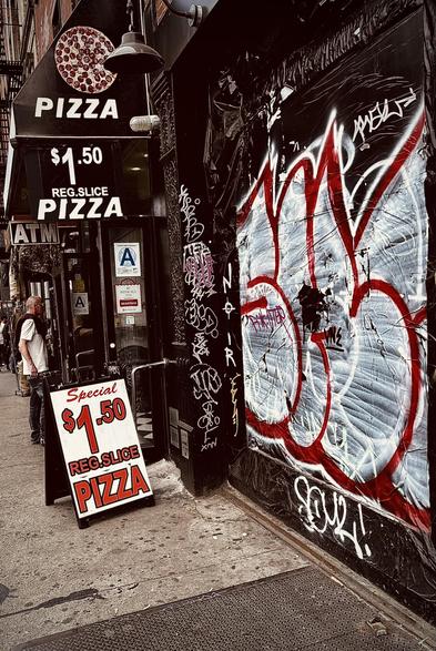 A color, wide-angle photograph in portrait orientation that captures a gritty, high-contrast street scene of a budget-friendly pizza shop on the Lower East Side of Manhattan. The shop's entrance is marked by multiple black signs and a sidewalk A-frame board advertising a "Special $1.50 Reg. Slice Pizza." Above the door, a black awning features a graphic of a pepperoni pizza and more bold white lettering. The storefront is narrow, with a visible "Grade A" health department rating in the window and an ATM sign protruding from the side. A man in a white t-shirt and glasses stands near the entrance, looking off-camera, while the sidewalk shows the typical wear and texture of a busy urban environment. The right side of the frame is dominated by a dark, textured wall heavily covered in street art. A large, stylized graffiti tag in bold red and white outlines sprawls across a corrugated metal shutter, layered over various smaller white and purple scrawls. The word "NOIR" is written vertically in white paint on a black pillar. The lighting is moody and dramatic, with deep shadows and desaturated tones that emphasize the urban decay and vibrant street culture. 