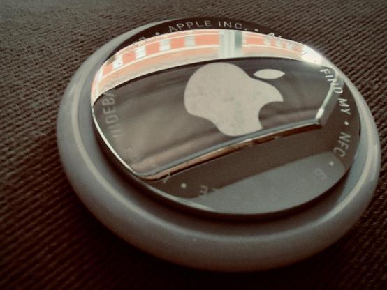A color, macro shot of an Apple AirTag resting on the textured, ribbed fabric surface of a vintage couch. The AirTag’s polished stainless steel battery cover occupies the center, reflecting an amber and white striped pattern of a nearby building through a window onto the AirTag. The Apple logo is prominently centered on the metallic disk, slightly scuffed from use. Encircling the metal edge, the engraved text reads "Apple Inc.," "AirTag," though some letters are obscured by the perspective and reflections. The image is treated with a warm, retro color grade, casting a sepia-like glow across the scene. The fabric beneath the device consists of thick, diagonal ridges in a deep chocolate brown and charcoal hue, emphasizing a tactile, mid-century aesthetic. The lighting is soft and directional, creating a gentle shadow beneath the rounded white plastic base of the AirTag, which makes the small tracking device appear nestled into the upholstery. The overall composition is tight and intimate, focusing on the contrast between the sleek, modern tech and the coarse, period-specific textile.