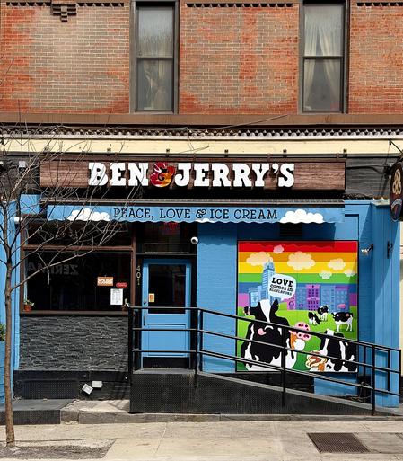 A color, wide-angle photograph that captures the street scene along 51st Street in the Hell’s Kitchen neighborhood of Manhattan. The focal point of the image is a colorful storefront of a Ben & Jerry’s ice cream shop, located at the base of a traditional red-brick building. A dark wood-textured sign with the brand’s name in bold white lettering and a colorful ampersand hangs above a blue awning that reads "PEACE, LOVE & ICE CREAM" in white. To the right of the blue-framed glass entrance door, a vibrant, large-scale mural features the iconic Ben & Jerry's cows in a grassy field against a rainbow-striped sky and a city skyline. A speech bubble in the mural declares, "LOVE COMES IN ALL FLAVORS." The lower half of the storefront is painted a bright sky blue, contrasting with a section of dark, textured slate-like stone under the large front window. A black metal handrail runs along a small ramp leading up to the entrance, which is marked with the street number 401. On the window, a small orange sign indicates the shop is currently closed. The scene is viewed from a straight-on perspective on the sidewalk, with a bare tree trunk visible on the far left and a metal utility grate set into the concrete in the foreground. Afternoon sunlight is evenly illumining the scene.