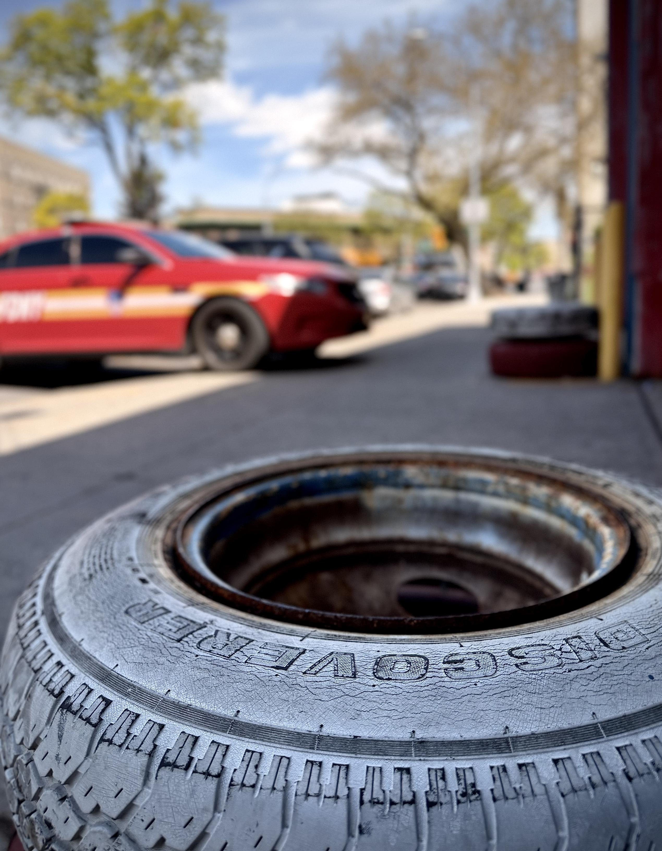 A color, wide-angle photograph in portrait orientation that captures the street scene along Boston Road in the Morrisania neighborhood of the Bronx. The focal point of the image is a close-up, low-angle shot featuring a weathered tire, resting on a concrete sidewalk in the foreground. The tire is a Cooper "Discoverer" model, with the brand name embossed clearly on its cracked and textured sidewall. The tire is mounted on a rusty metal rim, and it is beginning to flake, revealing the dark rubber beneath. This foreground element is in sharp focus, drawing immediate attention to the rugged, industrial texture of the rubber and the aging metal. In the soft-focus background, a vibrant red FDNY command vehicle is parked along the curb of a city street. Beyond the car, the blurred urban landscape of the Bronx unfolds under a bright blue sky with scattered clouds, including Spring time budding green trees and the distant brick facades of apartment buildings. The lighting is bright and natural, suggesting a clear spring afternoon, which creates a high-contrast scene between the tire and the saturated reds and greens of the city background.
