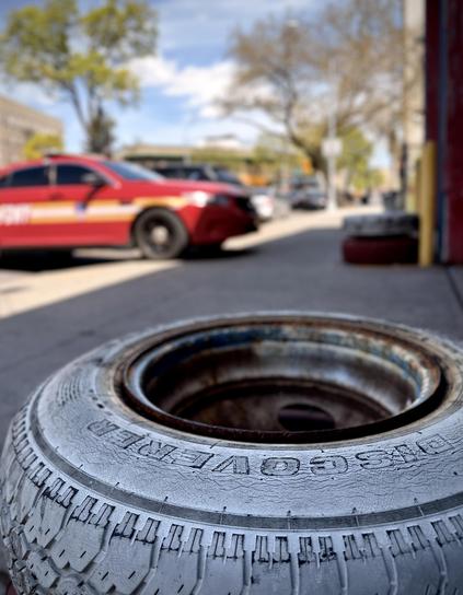A color, wide-angle photograph in portrait orientation that captures the street scene along Boston Road in the Morrisania neighborhood of the Bronx. The focal point of the image is a close-up, low-angle shot featuring a weathered tire, resting on a concrete sidewalk in the foreground. The tire is a Cooper "Discoverer" model, with the brand name embossed clearly on its cracked and textured sidewall. The tire is mounted on a rusty metal rim, and it is beginning to flake, revealing the dark rubber beneath. This foreground element is in sharp focus, drawing immediate attention to the rugged, industrial texture of the rubber and the aging metal. In the soft-focus background, a vibrant red FDNY command vehicle is parked along the curb of a city street. Beyond the car, the blurred urban landscape of the Bronx unfolds under a bright blue sky with scattered clouds, including Spring time budding green trees and the distant brick facades of apartment buildings. The lighting is bright and natural, suggesting a clear spring afternoon, which creates a high-contrast scene between the tire and the saturated reds and greens of the city background.