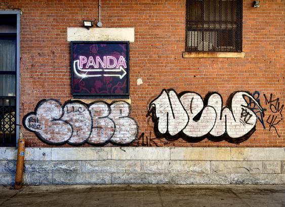 A color, wide-angle photograph in landscape orientation that captures the street scene along Riverside Drive in the Manhattanville neighborhood of Manhattan. The focal point of the scene is a weathered red brick wall of a multi-story building. Centered in the upper-left portion of the frame is a black-framed square lightbox displaying a glowing pink neon sign that reads "PANDA" in all caps. Below the text, a white neon arrow points to the right, finished with a stylized sparkle at its base. The sign is set against a dark purple background decorated with subtle floral patterns. A barred window with a dark screen sits recessed into the wall. The lower half of the brick facade is dominated by two large pieces of graffiti throw-ups. On the left, a rounded, bubble-style tag is filled with faded white paint and outlined in thick black. To its right, a sharper, more defined piece features bold white lettering with a heavy black drop shadow, accompanied by smaller black "tags" and the notation "4K" scrawled nearby. On the far left of the image, a vertical blue window frame is visible, partially obscured by a dark metal grate. A rusted orange bollard stands on the sidewalk in front of the wall, decorated with a few small, colorful stickers. The ground is a dark, damp concrete sidewalk that reflects afternoon sunlight.