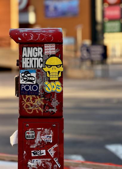 A color, wide-angle photograph in portrait orientation that captures the street scene along River Avenue in the Highbridge neighborhood of the Bronx, New York. The focal point of the image is a weathered red metal fire alarm call box standing on a street corner. The box is heavily decorated with various street art stickers and graffiti tags. Dominant stickers on the upper section include a large white "ANGR HEKTIC" decal, a stylized yellow skull with a mechanical jaw, and a blue and yellow "JOS" bubble-letter sticker. A blue "POLO HUE TECH" sticker and a smaller "I ❤️ MY HOOD" decal are also visible amidst various peeled and faded remnants of older labels. The red paint of the box is chipped and scratched, revealing silver metal underneath. The background is softly blurred with a shallow depth of field, looking across the intersection toward River Avenue and the Bronx Terminal Market. Out-of-focus elements include the tan and orange facades of large retail buildings, a dark blue billboard, and the white stripes of a pedestrian crosswalk on the asphalt. The warm morning sunlight casts soft shadows.
