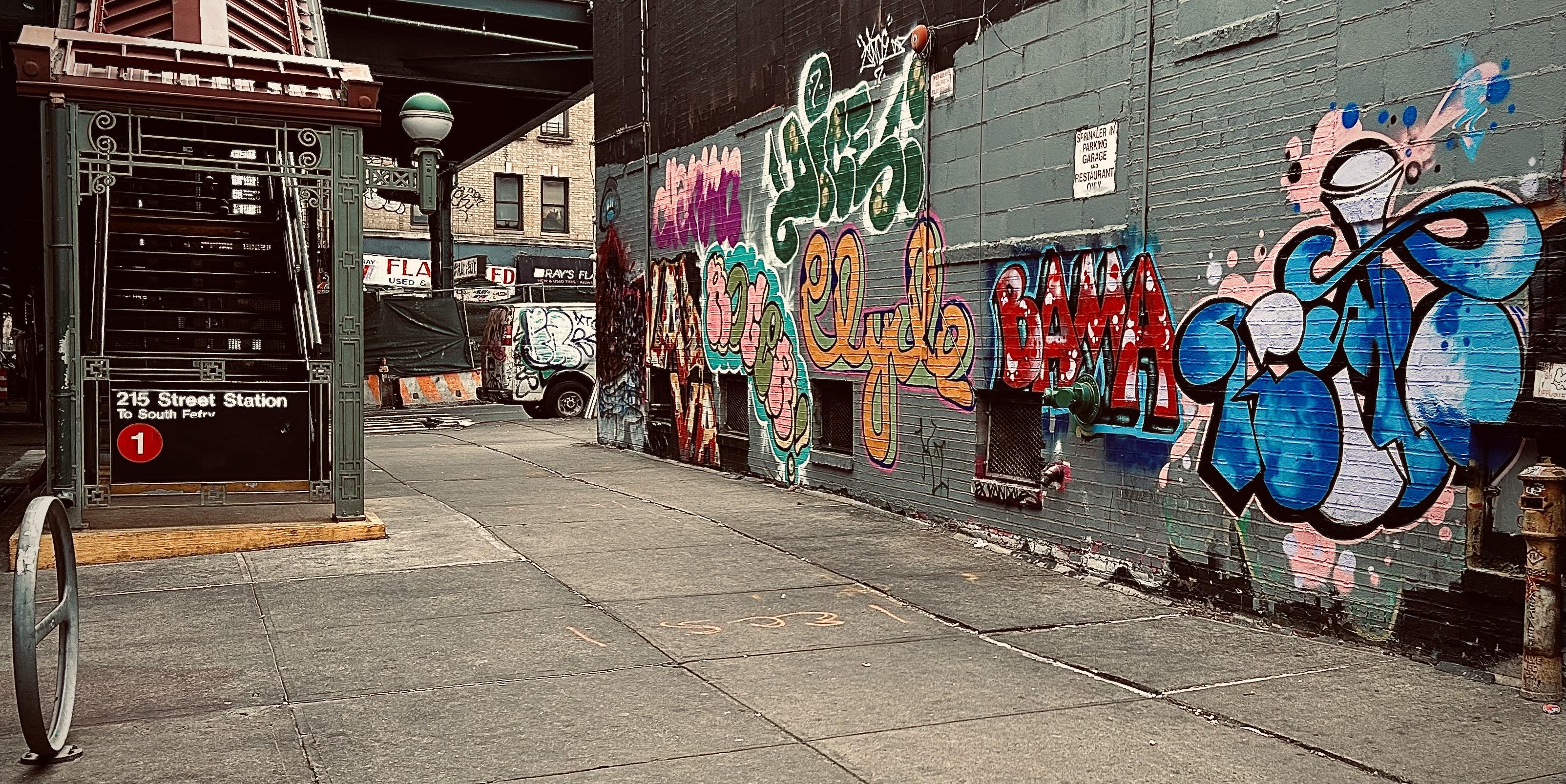 A color, wide-angle photograph in landscape orientation that captures the street scene along 10th Avenue in the Inwood neighborhood of Manhattan. The focal point of the image is a sidewalk flanked by a subway entrance and a heavily graffitied wall. On the left, the ornate green metal structure of the 215th Street Station subway entrance stairwell leads up to the elevated platform. A black sign with white text identifies it as "215 Street Station" for the "1" train, marked by a prominent red circle with a white "1" inside. In the background, a white van parked on the street is also covered in graffiti, and the dark underside of an elevated train track looms over the intersection. The right side of the image is dominated by several large, colorful graffiti pieces. The most prominent tags include a large "Clyde" in orange and yellow bubble letters, a bold red "BAMA" with white highlights, and a complex blue and white piece on the far right featuring swirling, liquid-like shapes. A small white sign warns that the area is a "Sprinkler in Parking Garage and Restaurant Only." The lighting is overcast and muted.