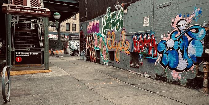 A color, wide-angle photograph in landscape orientation that captures the street scene along 10th Avenue in the Inwood neighborhood of Manhattan. The focal point of the image is a sidewalk flanked by a subway entrance and a heavily graffitied wall. On the left, the ornate green metal structure of the 215th Street Station subway entrance stairwell leads up to the elevated platform. A black sign with white text identifies it as "215 Street Station" for the "1" train, marked by a prominent red circle with a white "1" inside. In the background, a white van parked on the street is also covered in graffiti, and the dark underside of an elevated train track looms over the intersection. The right side of the image is dominated by several large, colorful graffiti pieces. The most prominent tags include a large "Clyde" in orange and yellow bubble letters, a bold red "BAMA" with white highlights, and a complex blue and white piece on the far right featuring swirling, liquid-like shapes. A small white sign warns that the area is a "Sprinkler in Parking Garage and Restaurant Only." The lighting is overcast and muted.