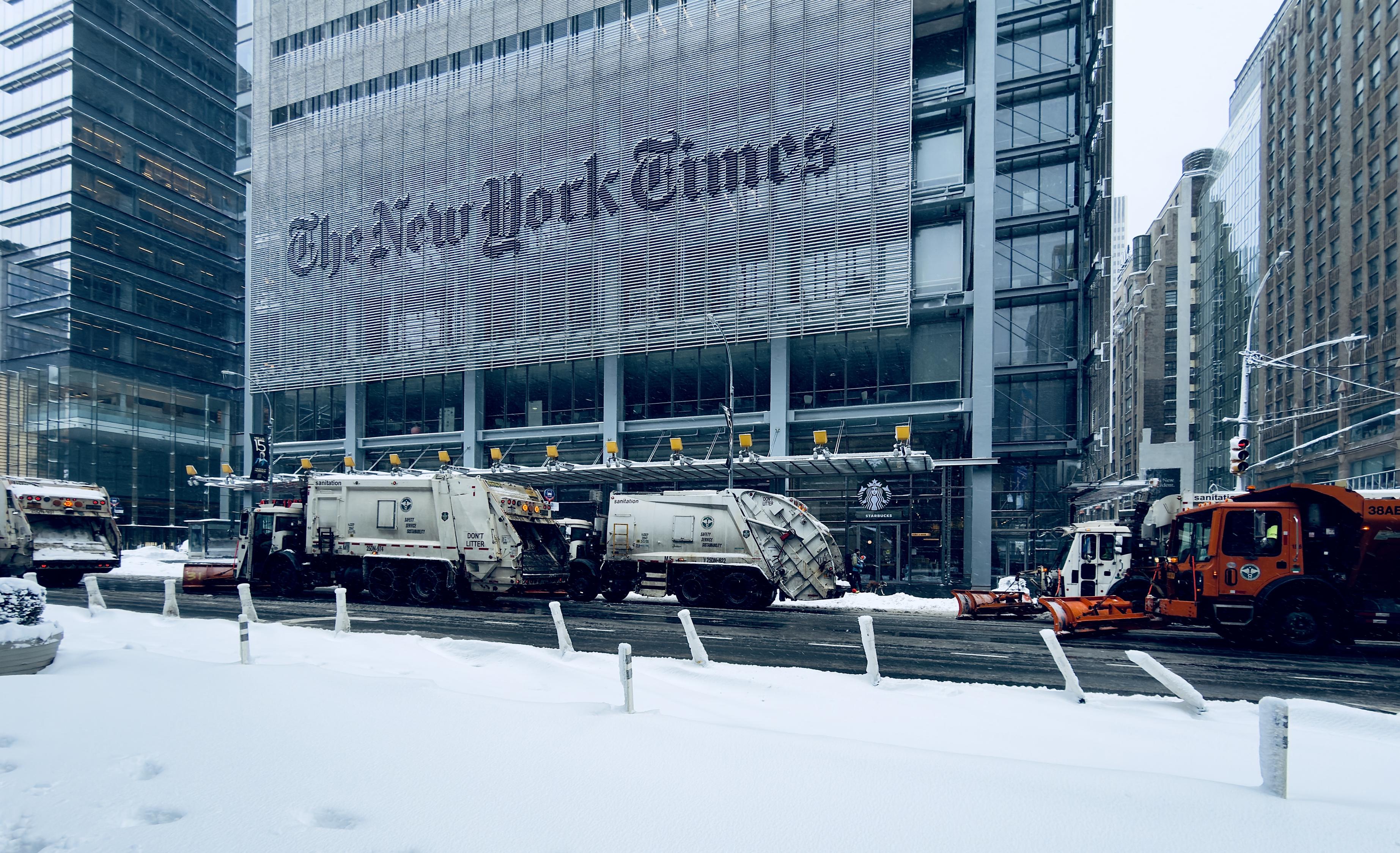A color, wide-angle photograph in landscape orientation that captures a fleet of Department of Sanitation trucks lined up along a snow-covered Eighth Avenue in Manhattan. In the background, the distinctive glass and steel facade of the New York Times Building dominates the frame, featuring the iconic "The New York Times" masthead across its sunshade screen. A Starbucks storefront is visible at the base of the skyscraper. The street in the foreground is heavily blanketed in white snow, with small white bollards partially buried along the edge of the roadway. The surrounding urban landscape is filled with modern high-rise buildings and classic brick structures, their details slightly softened by the hazy, grey light of the snowy day.