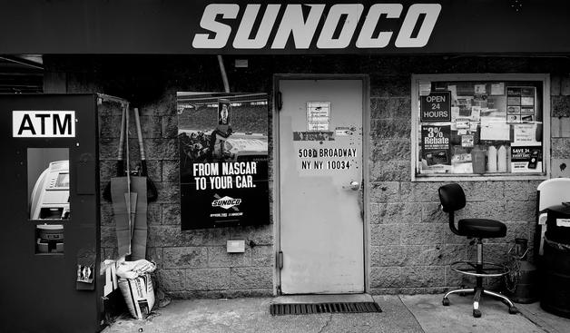 A noir, wide-angle photograph in landscape orientation that captures a gritty, functional storefront of a Sunoco station along Broadway in the Inwood neighborhood of Manhattan. The composition is dominated by a textured cinderblock wall and a heavy metal door at the center, flanked by an outdoor ATM kiosk on the left and a cluttered service window on the right. A large, bold "SUNOCO" sign hangs across the top of the frame, casting a slight shadow over the scene. To the left of the door, a NASCAR promotional poster titled "FROM NASCAR TO YOUR CAR" is plastered on the wall, and various tools, including long-handled shovels and a bag of ice melt, lean against the structure. A lone, black adjustable swivel chair with a backrest sits on the concrete sidewalk in front of the window.