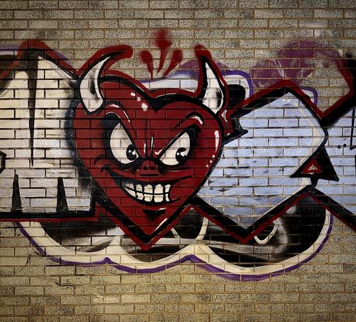 A color, medium-angle photograph in square aspect ratio that captures a vibrant piece of street art painted onto a light-colored, tan brick wall along Walton Avenue in the Highbridge neighborhood of the Bronx. The central focus is a large, stylized red heart depicted with a mischievous, "devilish" persona. The heart features two white, curved horns protruding from its upper lobes and a face with large, expressive white eyes, furrowed black eyebrows, and a wide, toothy grin that reveals a row of clenched teeth. The character has a small pig-like nose and subtle white highlights that give the red surface a glossy, three-dimensional appearance. The character is integrated into a larger graffiti composition, flanked by bold, geometric lettering. The lighting is warm and slightly dramatic, casting soft shadows that emphasize the grit and texture of the urban canvas.