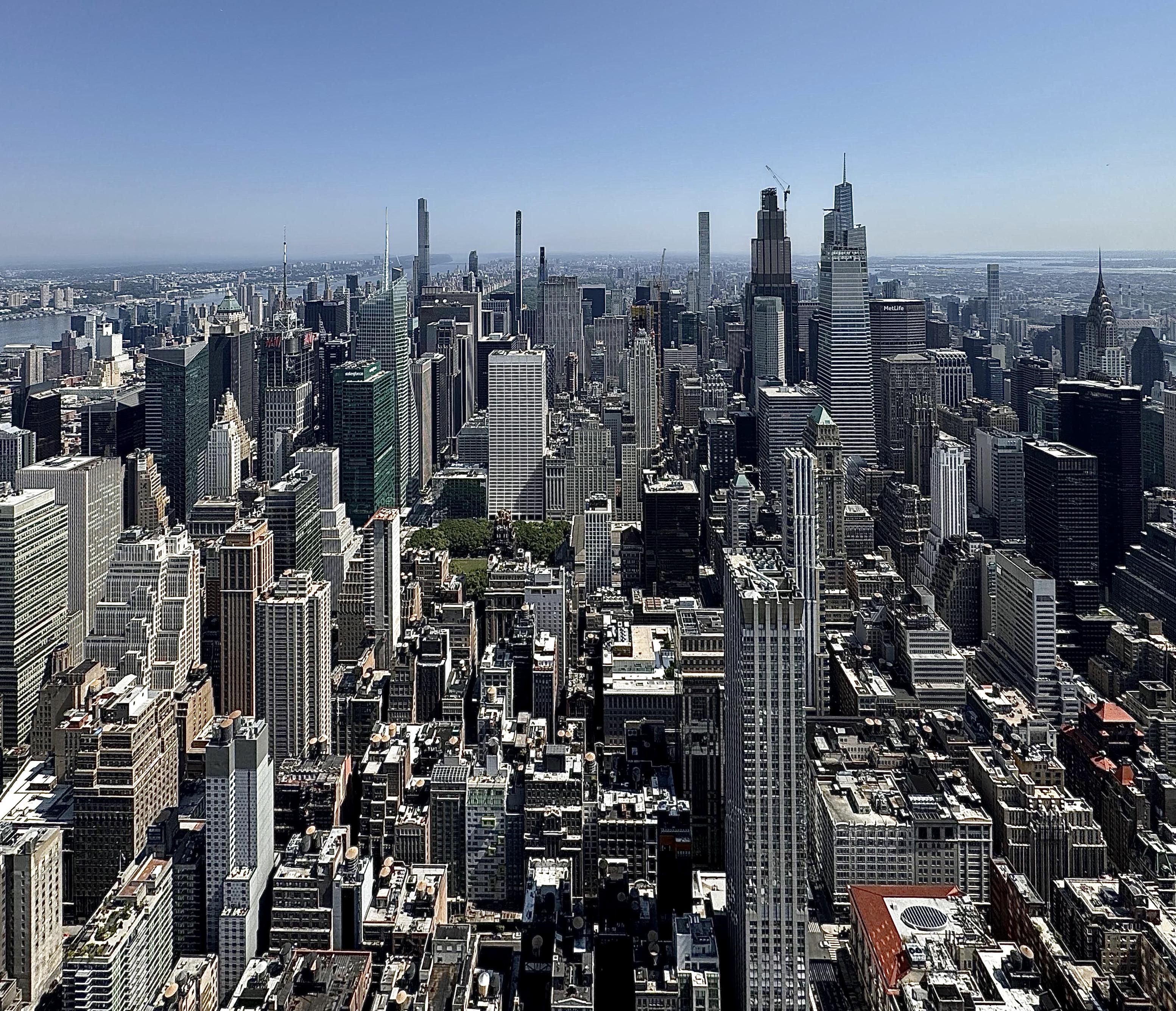 A color, wide-angle landscape photograph in square aspect ratio that captures the dense urban landscape of Midtown Manhattan on. The view is looking north from the Empire State Building under a clear, bright blue sky. In the immediate foreground, a chaotic yet rhythmic collection of rooftops, water towers, and smaller office buildings creates a textured foundation of tan, gray, and brick-red tones. Moving into the mid-ground, the greenery of Bryant Park offers a small bit of color amidst the concrete. To the right, the iconic art deco spire of the Chrysler Building stands out, while the massive, modern glass facades of the Bank of America Tower and the Salesforce Tower dominate the center-left. To the far left, the Hudson River is visible as a shimmering blue strip, separating the dense grid of Manhattan from the hazy coastline of New Jersey. The lighting is sharp and direct, casting deep shadows that emphasize the geometric depth of the city's "canyons."