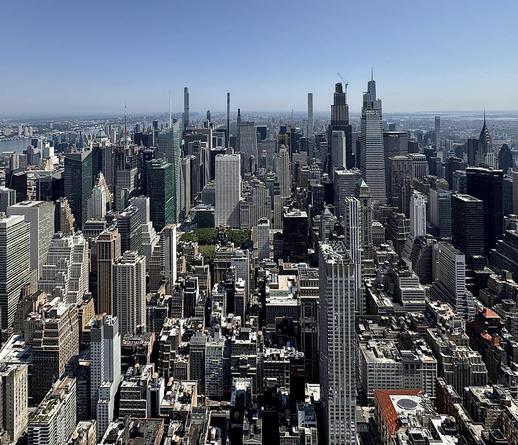 A color, wide-angle landscape photograph in square aspect ratio that captures the dense urban landscape of Midtown Manhattan on. The view is looking north from the Empire State Building under a clear, bright blue sky. In the immediate foreground, a chaotic yet rhythmic collection of rooftops, water towers, and smaller office buildings creates a textured foundation of tan, gray, and brick-red tones. Moving into the mid-ground, the greenery of Bryant Park offers a small bit of color amidst the concrete. To the right, the iconic art deco spire of the Chrysler Building stands out, while the massive, modern glass facades of the Bank of America Tower and the Salesforce Tower dominate the center-left. To the far left, the Hudson River is visible as a shimmering blue strip, separating the dense grid of Manhattan from the hazy coastline of New Jersey. The lighting is sharp and direct, casting deep shadows that emphasize the geometric depth of the city's "canyons."
