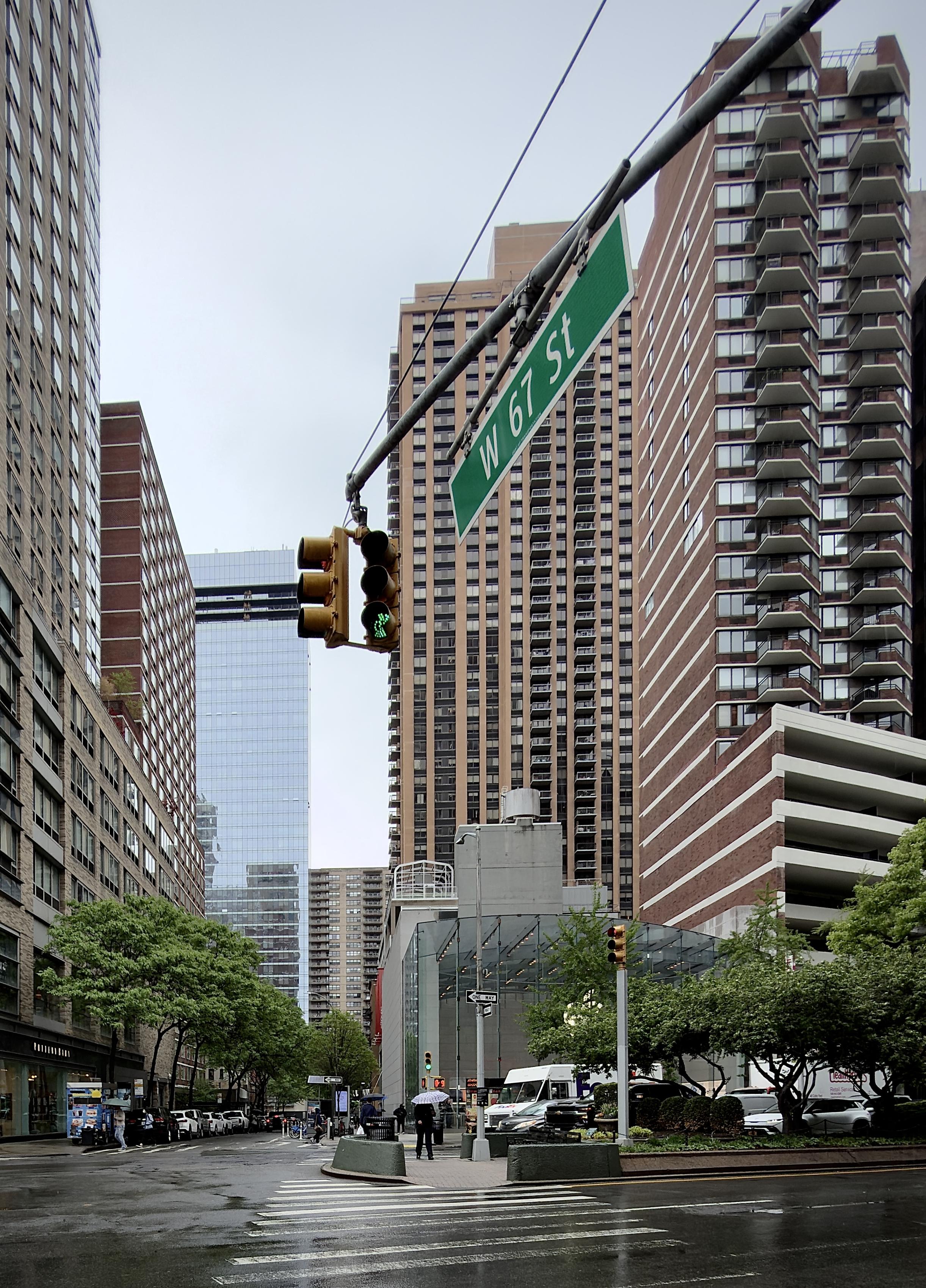 A color, wide-angle photograph in portrait orientation that captures the intersection of Broadway and West 67th Street on the Upper West Side of Manhattan during a rainy, gloomy, overcast day. The wet asphalt of the crosswalk in the foreground reflects the flat, gray light of the sky. Dominating the upper center of the frame is a green street sign for "W 67 St" angled over the street, hanging from a metal arm alongside a vertical traffic signal showing a green light. In the background, the modern, glass-facade Apple Upper West Side store sits at the base of a towering, tan-colored residential skyscraper with a repetitive grid of windows and balconies. Several pedestrians carrying umbrellas are visible near the Apple Store’s glass entrance, and a white FedEx delivery truck is parked at the curb. Lush green trees line the sidewalks. 
