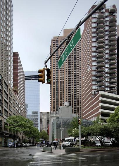 A color, wide-angle photograph in portrait orientation that captures the intersection of Broadway and West 67th Street on the Upper West Side of Manhattan during a rainy, gloomy, overcast day. The wet asphalt of the crosswalk in the foreground reflects the flat, gray light of the sky. Dominating the upper center of the frame is a green street sign for "W 67 St" angled over the street, hanging from a metal arm alongside a vertical traffic signal showing a green light. In the background, the modern, glass-facade Apple Upper West Side store sits at the base of a towering, tan-colored residential skyscraper with a repetitive grid of windows and balconies. Several pedestrians carrying umbrellas are visible near the Apple Store’s glass entrance, and a white FedEx delivery truck is parked at the curb. Lush green trees line the sidewalks. 