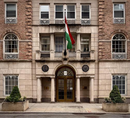 A color, wide-angle photograph in square aspect ratio that captures the symmetrical, neoclassical facade of the Consulate General of Hungary in Manhattan. The building is constructed from a mix of light-colored limestone on the lower levels and reddish-brown brick on the upper floors, featuring ornate white wrought-iron window guards throughout. A large Hungarian flag hangs prominently from a central flagpole above the entrance. On either side of the main archway, two dark circular medallions are mounted on the stone; the left displays the Hungarian coat of arms, while the right features the emblem of the United Nations. The sidewalk in front is flanked by two large stone planters containing manicured evergreen shrubs and small flowers, grounding the stately architecture. The central entrance is defined by a grand stone archway supported by four simple, elegant columns. Set within this arch are the double doors, which are crafted from a warm, polished wood with a golden-brown finish. Each door features a large, multi-paned glass window that allows a view into the interior, echoing the grid pattern of the building’s upper windows. A small white security notice is visible on the right-hand door.