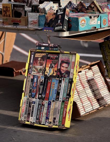 A color, wide-angle photograph in portrait orientation that captures a street vendor’s eclectic display of vintage physical media on a sidewalk at Union Square in Manhattan. A white folding table serves as the primary platform, cluttered with a variety of VHS tapes and CDs housed in repurposed cardboard fruit crates, such as blue "Sunset" produce boxes. On the table, notable titles include a Nelly Furtado CD and VHS copies of The Godfather and Rider on the Rain. The background is softly blurred, showing a brown briefcase and the sun-drenched pavement. On the ground, leaning against the table leg, a bright yellow "Chiquita" banana box is repurposed as a vertical display case for an array of VHS tapes. The top row features prominent titles like Sahara starring Humphrey Bogart, Beverly Hills Cop II, and an Elvis Presley documentary. Below them, two densely packed rows of tapes showcase a mix of genres. To the right, another cardboard tray lies angled on the ground, filled with rows of compact discs in jewel cases, their white spines showing.