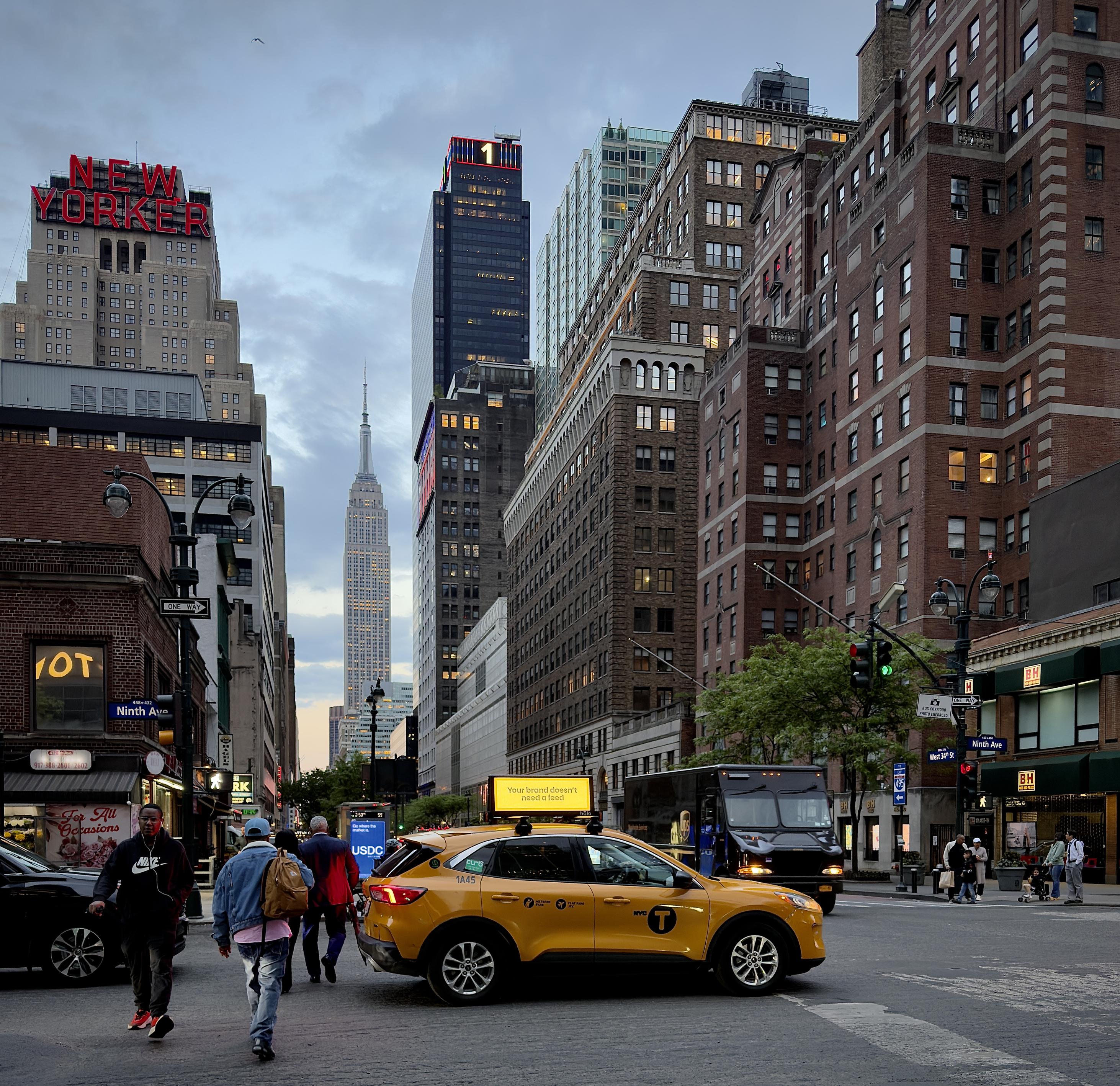 A color, wide-angle photograph in square aspect ratio that captures the street scene along Ninth Avenue in the Chelsea neighborhood of Manhattan, during dusk. In the center foreground, a bright yellow taxi SUV is positioned as if stopped in traffic, its yellow "NYC Taxi" emblem and an advertisement on its roof clearly visible that reads “Your brand doesn’t need a feed.” To its right, a dark UPS delivery truck is partially visible, while on the left, several pedestrians are captured mid-stride crossing the street. The composition utilizes the city’s "urban canyon" effect, drawing the eye straight down the street to the Empire State Building, which stands illuminated in a pale, silvery glow against the darkening sky in the far distance. On the far left, the massive red neon "NEW YORKER" sign sits atop the New Yorker Hotel, and a modern, dark skyscraper nearby features a bright red neon numeral "1" at its summit. To the right, tall, classic brick apartment buildings rise above the street, with the recognizable green awnings of a B&H Photo Video store marking the corner. The sky is a muted, overcast blue-grey, typical of early evening.
