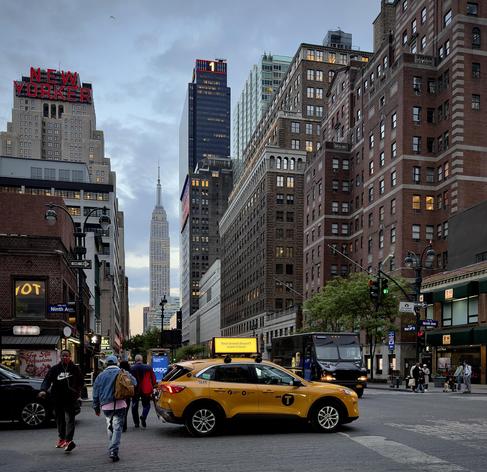 A color, wide-angle photograph in square aspect ratio that captures the street scene along Ninth Avenue in the Chelsea neighborhood of Manhattan, during dusk. In the center foreground, a bright yellow taxi SUV is positioned as if stopped in traffic, its yellow "NYC Taxi" emblem and an advertisement on its roof clearly visible that reads “Your brand doesn’t need a feed.” To its right, a dark UPS delivery truck is partially visible, while on the left, several pedestrians are captured mid-stride crossing the street. The composition utilizes the city’s "urban canyon" effect, drawing the eye straight down the street to the Empire State Building, which stands illuminated in a pale, silvery glow against the darkening sky in the far distance. On the far left, the massive red neon "NEW YORKER" sign sits atop the New Yorker Hotel, and a modern, dark skyscraper nearby features a bright red neon numeral "1" at its summit. To the right, tall, classic brick apartment buildings rise above the street, with the recognizable green awnings of a B&H Photo Video store marking the corner. The sky is a muted, overcast blue-grey, typical of early evening.