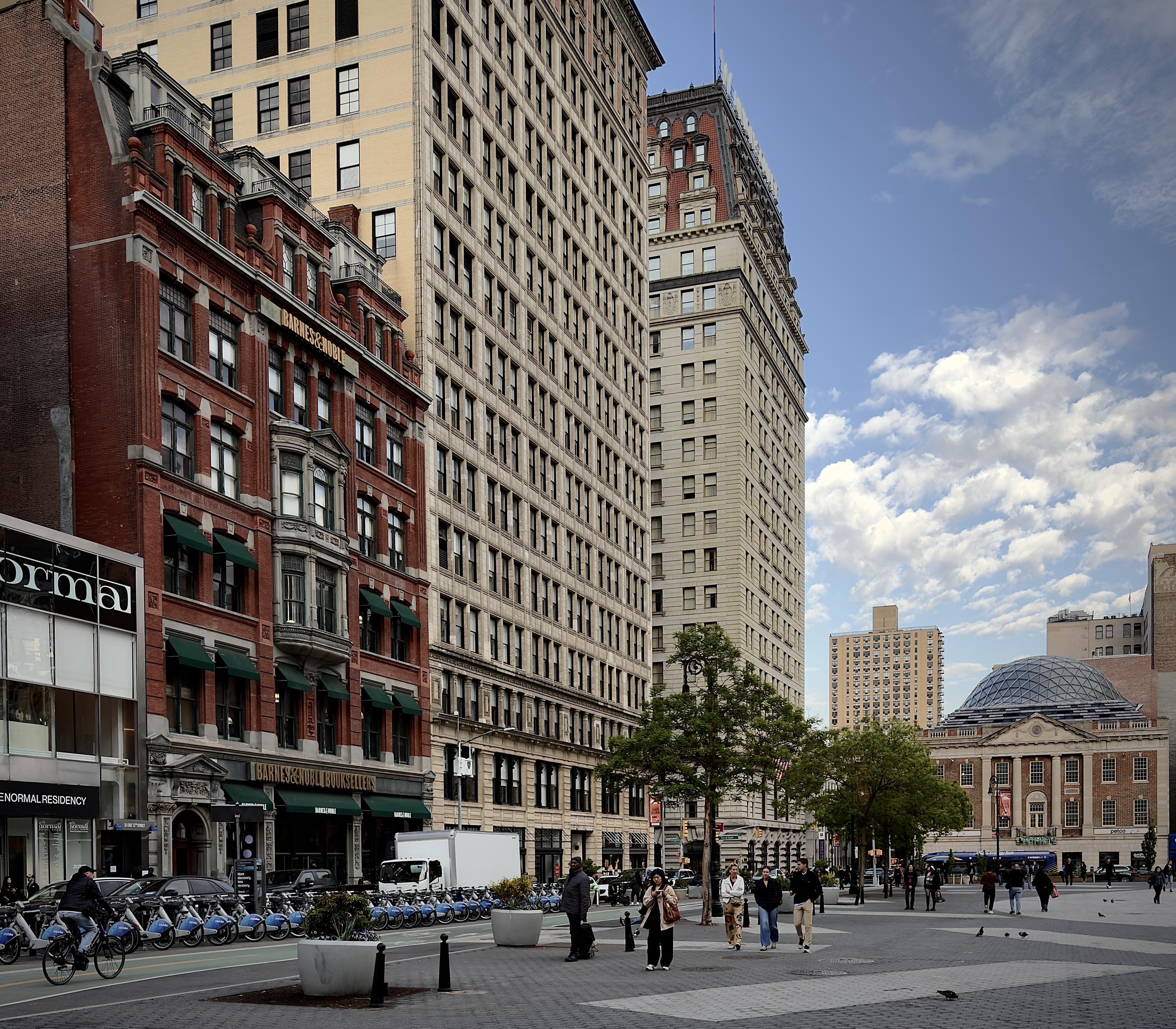 A color, wide-angle photograph in square aspect ratio that captures the street scene of Union Square in Manhattan under a bright blue sky filled with scattered, wispy white clouds. On the left, a grand five-story building of deep terracotta brick features ornate stone carvings and several rows of windows shaded by forest-green fabric awnings. A Barnes & Noble bookstore, this structure stands in stark contrast to the towering neoclassical skyscraper immediately beside it, which rises many stories high in a pale parchment-colored stone with a rhythmic grid of uniform windows. Further down the street, a distinct building with a charcoal-toned mansard roof punctuates the skyline. In the foreground, a wide pedestrian plaza paved in slate-gray stone is populated by several people walking in various directions. A cyclist in dark clothing pedals across the lower left, passing a long, orderly row of sapphire-blue shared bicycles docked along the curb.