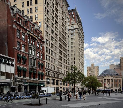 A color, wide-angle photograph in square aspect ratio that captures the street scene of Union Square in Manhattan under a bright blue sky filled with scattered, wispy white clouds. On the left, a grand five-story building of deep terracotta brick features ornate stone carvings and several rows of windows shaded by forest-green fabric awnings. A Barnes & Noble bookstore, this structure stands in stark contrast to the towering neoclassical skyscraper immediately beside it, which rises many stories high in a pale parchment-colored stone with a rhythmic grid of uniform windows. Further down the street, a distinct building with a charcoal-toned mansard roof punctuates the skyline. In the foreground, a wide pedestrian plaza paved in slate-gray stone is populated by several people walking in various directions. A cyclist in dark clothing pedals across the lower left, passing a long, orderly row of sapphire-blue shared bicycles docked along the curb.