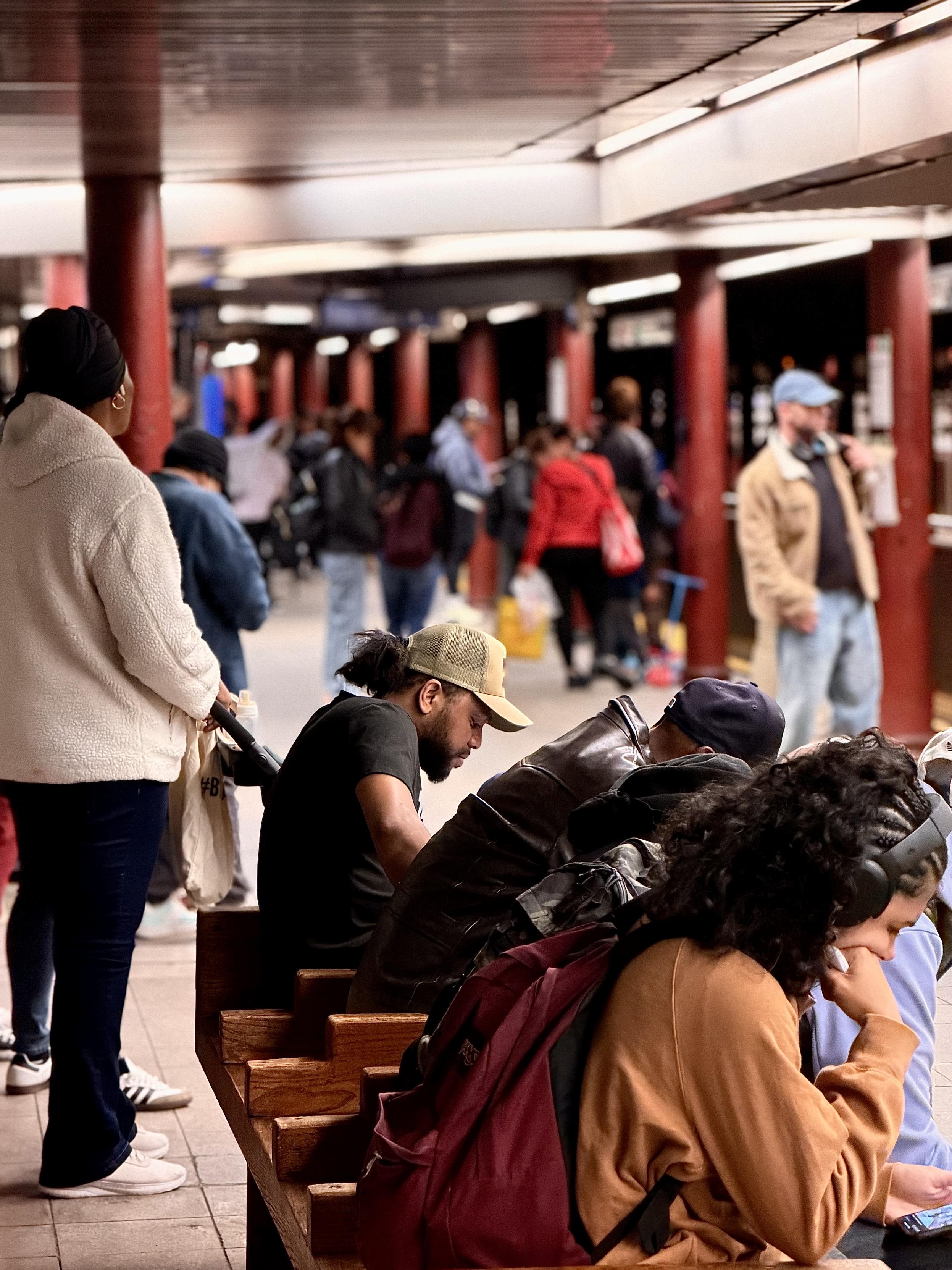 A color, wide-angle photograph in portrait orientation that captures the scene on a Manhattan subway platform. In the foreground, several individuals are seated on a wooden bench, mostly looking down at their phones or lost in thought. A man with a tan baseball cap and a black t-shirt sits near the center, while a person with long, dark curly hair and over-ear headphones occupies the right foreground, wearing an orange sweatshirt and carrying a maroon backpack. To the left, a woman stands with her back to the camera, wearing a white fleece jacket, dark pants, and a black head covering, holding the handle of a stroller or shopping cart. The background reveals the deep perspective of the long subway station, lined with thick, dark red columns that recede into a soft blur. Numerous commuters are scattered along the platform, some standing near the tracks and others walking through the space. The lighting is artificial and slightly dim, typical of an underground station, with bright overhead lights reflecting off the smooth, light-colored floor tiles.