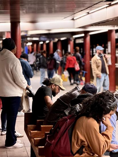 A color, wide-angle photograph in portrait orientation that captures the scene on a Manhattan subway platform. In the foreground, several individuals are seated on a wooden bench, mostly looking down at their phones or lost in thought. A man with a tan baseball cap and a black t-shirt sits near the center, while a person with long, dark curly hair and over-ear headphones occupies the right foreground, wearing an orange sweatshirt and carrying a maroon backpack. To the left, a woman stands with her back to the camera, wearing a white fleece jacket, dark pants, and a black head covering, holding the handle of a stroller or shopping cart. The background reveals the deep perspective of the long subway station, lined with thick, dark red columns that recede into a soft blur. Numerous commuters are scattered along the platform, some standing near the tracks and others walking through the space. The lighting is artificial and slightly dim, typical of an underground station, with bright overhead lights reflecting off the smooth, light-colored floor tiles.