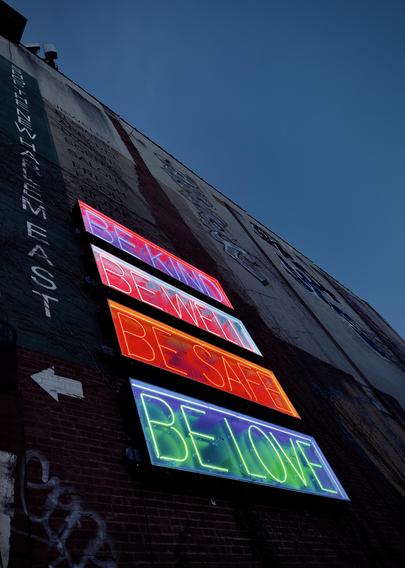 A color, wide-angle photograph in portrait orientation that captures a tall brick wall along Fifth Avenue in the East Harlem neighborhood of Manhattan near sunset. The wall rises into a deep blue twilight sky, showcasing a blend of commercial history and modern street art. Four large, rectangular neon signs are mounted vertically on the wall, glowing brilliantly against the darkening brick. From top to bottom, the signs display the phrases "BE KIND" in pink, "BE WELL" in white, "BE SAFE" in bright orange, and "BE LOVE" in vibrant green. The neon light reflects slightly against the inner frames of the boxes, creating a saturated, luminous effect that contrasts sharply with the weathered textures of the building. To the left, a vertical green banner features white lettering that reads "SHOP THE NEW HARLEM EAST." Above and around the neon signs, the wall is covered in layers of faded painted advertisements for jewelry and salons, alongside large-scale grey and black graffiti tags. A white arrow points toward the left on the lower brick portion, while delicate, dormant vines creep up the surface.