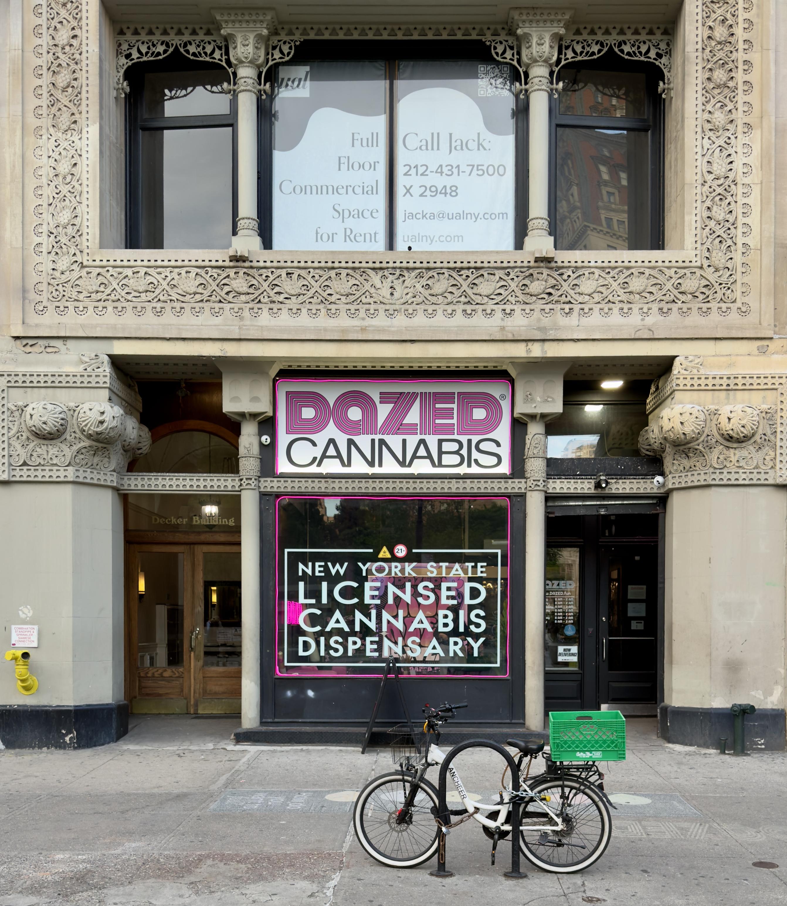 A color, wide-angle photograph that captures the exterior of a cannabis dispensary in Union Square, Manhattan. A historic, ornate stone building with intricate floral carvings and decorative columns surrounds the storefront. Centered in the frame is a large window and signage for "DAZED CANNABIS," featuring a bold, pink-and-white stylized logo at the top. Below the logo, a large window is bordered by a bright pink neon light, displaying text that identifies the business as a "NEW YORK STATE LICENSED CANNABIS DISPENSARY." To the left of this central display is a set of arched, wooden double doors labeled "Decker Building," while to the right is a sleek, black modern door that serves as the primary entrance for the dispensary. On the sidewalk in front of the building, a white electric bicycle with a green plastic crate attached to the rear rack is locked to a circular metal bike rack. The building's architecture above the storefront includes a balcony with a stone railing and large windows that feature a "Full Floor Commercial Space for Rent" sign.