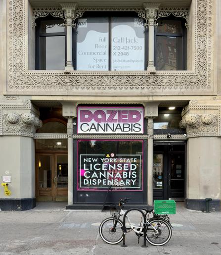 A color, wide-angle photograph that captures the exterior of a cannabis dispensary in Union Square, Manhattan. A historic, ornate stone building with intricate floral carvings and decorative columns surrounds the storefront. Centered in the frame is a large window and signage for "DAZED CANNABIS," featuring a bold, pink-and-white stylized logo at the top. Below the logo, a large window is bordered by a bright pink neon light, displaying text that identifies the business as a "NEW YORK STATE LICENSED CANNABIS DISPENSARY." To the left of this central display is a set of arched, wooden double doors labeled "Decker Building," while to the right is a sleek, black modern door that serves as the primary entrance for the dispensary. On the sidewalk in front of the building, a white electric bicycle with a green plastic crate attached to the rear rack is locked to a circular metal bike rack. The building's architecture above the storefront includes a balcony with a stone railing and large windows that feature a "Full Floor Commercial Space for Rent" sign.