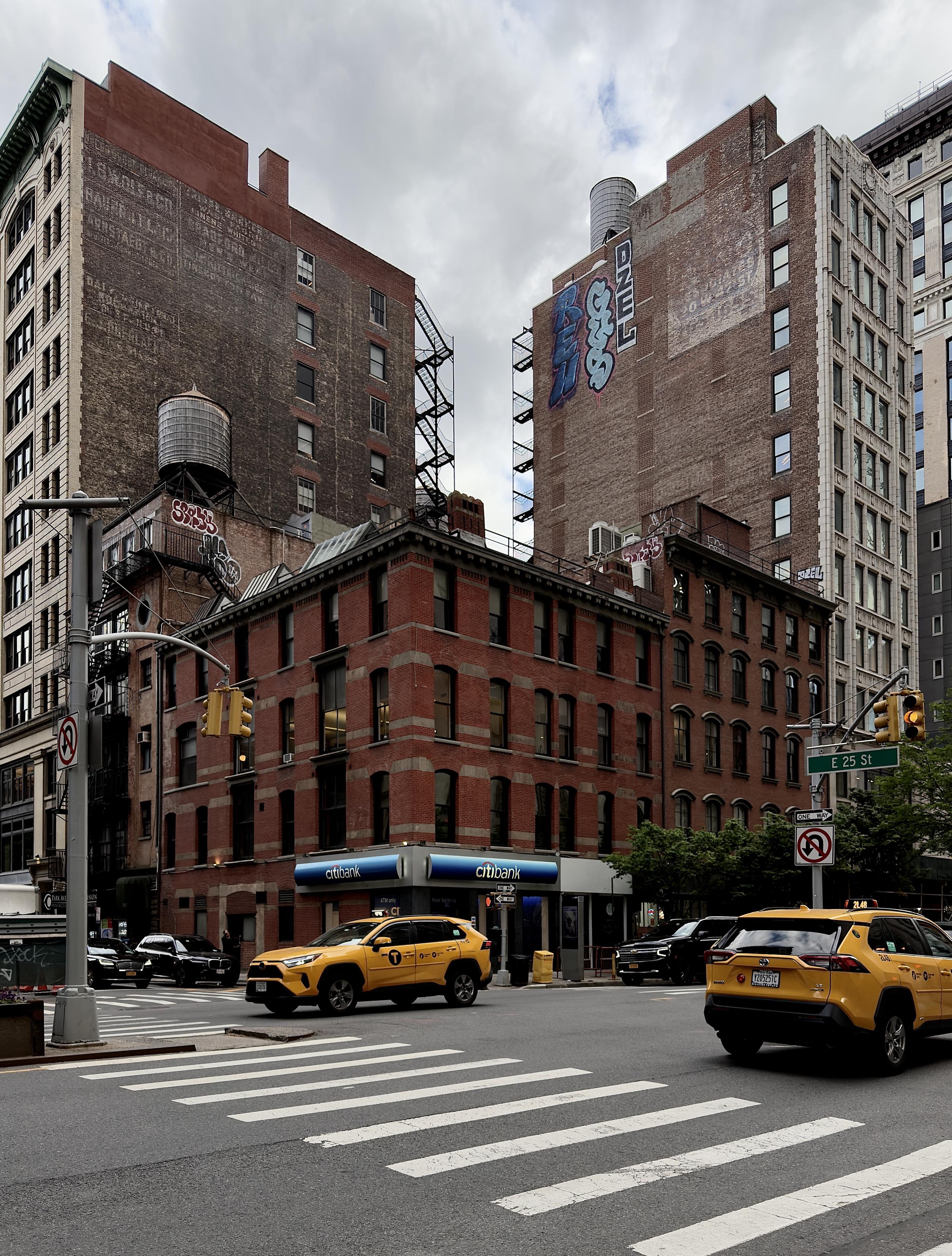 A color, wide-angle photograph in portrait orientation that captures the street scene along Park Avenue South in the Rose Hill neighborhood of Manhattan under an overcast, cloudy afternoon sky. In the foreground, a prominent zebra crosswalk stretches across the asphalt road, where two yellow NYC taxi SUVs are captured in mid-motion driving in opposite directions along the avenue. The corner is anchored by a four-story, red brick commercial building featuring a Citibank branch on its ground floor with its signature blue signage. Above it, towering brick apartment and office buildings rise into the background, showcasing classic architectural details like black fire escapes, arched windows, and traditional wooden water towers perched on the roofs. Bold, colorful graffiti tags, including one reading "DZEL," are sprayed high up on the exposed side walls. Street infrastructure frames the intersection, including a silver traffic light pole with a "No Right Turn" sign on the left, and a green street sign marking "E 25 St" on the right. Several dark-colored cars and SUVs are parked or idling in the background near the cross streets.