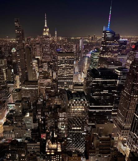 A color, wide-angle, high-angle photograph in landscape orientation captured at nighttime. A sweeping view of the Midtown and Lower Manhattan skyline looking south from the Top of the Rock observation deck at Rockefeller. The Empire State Building stands prominently in the upper-left center, its upper tiers and towering spire brilliantly illuminated in bright white light against a pitch-black sky. Far in the background, near the horizon line, the glowing silhouette and spire of One World Trade Center are faintly visible. The surrounding cityscape is a dense, geometric tapestry of skyscrapers stretching toward the horizon, filled with thousands of glowing yellow and white windows that create a glittering grid pattern. In the foreground and midground, towering structures of various architectural styles block and layer over one another, forming deep, dark street canyons where faint street-level illumination peeks through.