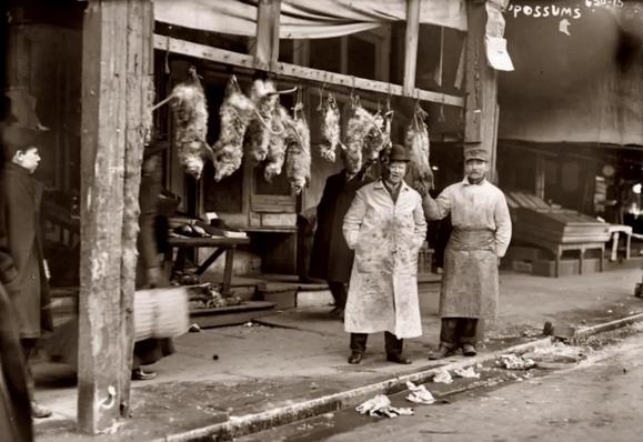 A sepia-toned, wide-angle photograph in landscape orientation, captured in 1915, that depicts the street scene along the Bowery in New York City. The photograph features a market stall specializing in selling opossums. In the foreground, two men stand side-by-side; the man on the left wears a light-colored, blood-stained apron over his coat and a brimmed hat, while the man to his right is dressed in a dark uniform and cap, wearing an apron as well. Behind them, a wooden rack suspended from the storefront’s overhang holds several opossums hanging by their tails, clearly intended for sale. A sign at the top right of the frame explicitly labels the establishment's offerings with the word "OPOSSUMS,". To the far left, a young boy stands observing the scene, partially obscured by a large wooden support beam that frames that side of the image. Another figure, partially blurred, walks past the stall. The ground is littered with scattered debris.