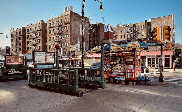 A color, wide-angle photograph in landscape orientation that captures a sunny, late afternoon street scene outside the entrance of a subway station along Dyckman Street in the Inwood neighborhood of Manhattan. In the foreground, a prominent green metal railing encloses the stairs descending into the subway. To the right of the entrance stands a vibrant red food cart topped with yellow and blue "Sabrett" umbrellas, advertising an array of hot dogs, sandwiches, and drinks. The cart is positioned on the sidewalk, with a large, yellow traffic light structure looming nearby. The proprietor stands behind the counter. In the background, a row of fire escape clad multi-story brick apartment buildings line the street. A McDonald’s sign is visible further down the street, and the overall atmosphere is one of a bustling urban transit point, with advertisements posted on the subway structure and clear shadows indicating the low angle of the sun.