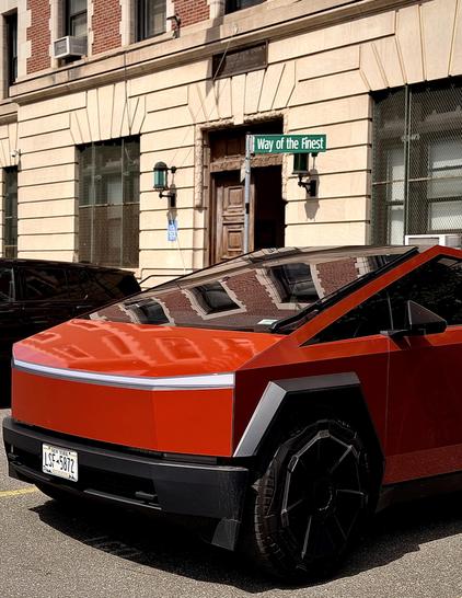 A color, wide-angle photograph in portrait orientation that captures the street scene along Third Avenue in the Melrose neighborhood of the Bronx. The focal point of the place is a Tesla Cybertruck in a custom, vibrant orange color parked on an asphalt street in front of a public office. The vehicle’s angular, stainless-steel form is accentuated by the bright finish, which starkly contrasts with its matte black wheel arches, large black wheels, and the dark trim along the lower bumper. There is a New York license plate clearly visible on the front bumper. In the background, a building identified by the signage "42nd Precinct" and a street sign reading "Way of the Finest" provides a clear sense of place. The scene is captured in clear daylight, with shadows cast by the building and the vehicle, while the reflective glass of the Cybertruck’s panoramic windshield mirrors the surrounding architecture.