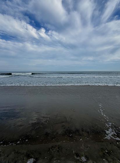 A color, wide-angle photograph in portrait orientation that captures a quiet, expansive stretch of the beach in Atlantic City, New Jersey, under a mostly cloudy sky afternoon sky. The horizon line is positioned in the upper third of the frame, where the deep blue Atlantic Ocean meets a complex sky filled with wispy, streaky cloud formations that blend patches of clear blue with soft, overcast white and grey tones. A gentle surf with small, rhythmic waves rolls toward the shoreline, creating a thin, white foamy edge that contrasts against the dark, wet sand in the foreground. The beach itself appears wide and flat, with the sand darkened by recent moisture, reflecting the soft, diffused light from above. There are no people, structures, or debris visible, emphasizing the vastness of the shoreline.