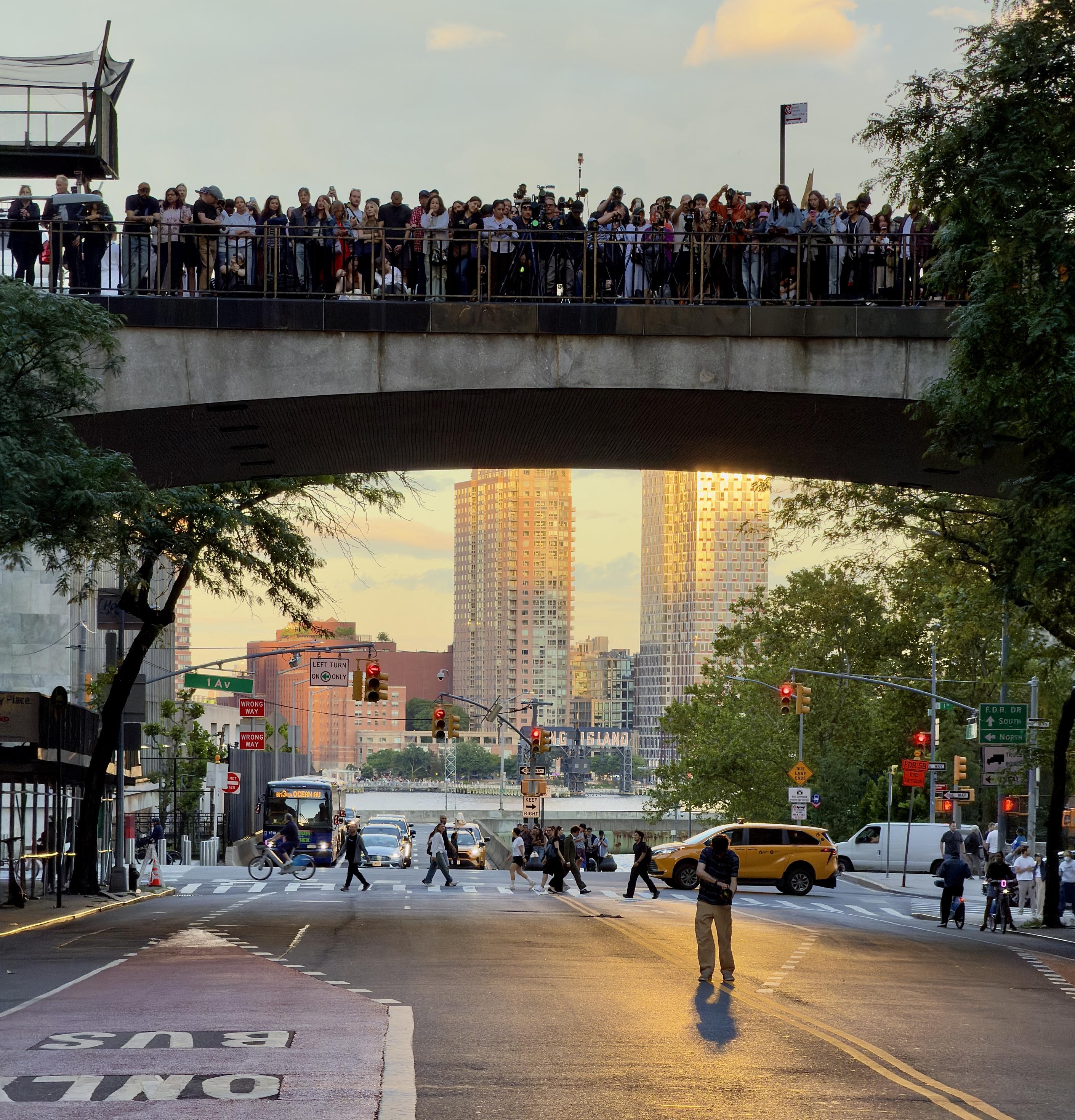 A color, wide-angle photograph in square aspect ratio that captures a large crowd of people lining the elevated Tudor City pedestrian bridge, silhouetted against the bright evening sky as they wait for the Manhattanhenge sunset in New York City. Many individuals in the crowd are holding cameras with long lenses or smartphones, aimed westward toward the street level, capturing the golden light reflecting off the tall buildings in the background. The bridge spans across the upper portion of the frame, creating a dark, wide archway that frames a view of the street below, where the warm, low-angled sunlight illuminates the pavement and surrounding structures. Below the bridge in "image.png," the street scene is active, with pedestrians walking across the crosswalk and a yellow taxi navigating the intersection. Traffic lights are visible, casting a glow over the asphalt, which shows long, soft shadows stretching toward the viewer, characteristic of the minutes before sunset. To the left, a "BUS ONLY" lane is painted on the road, and trees frame the edges of the composition, leading the eye toward the center where the sunlit skyscrapers glisten against a pale, clear sky. A green street sign reads 1 Av, hanging from a traffic pole.