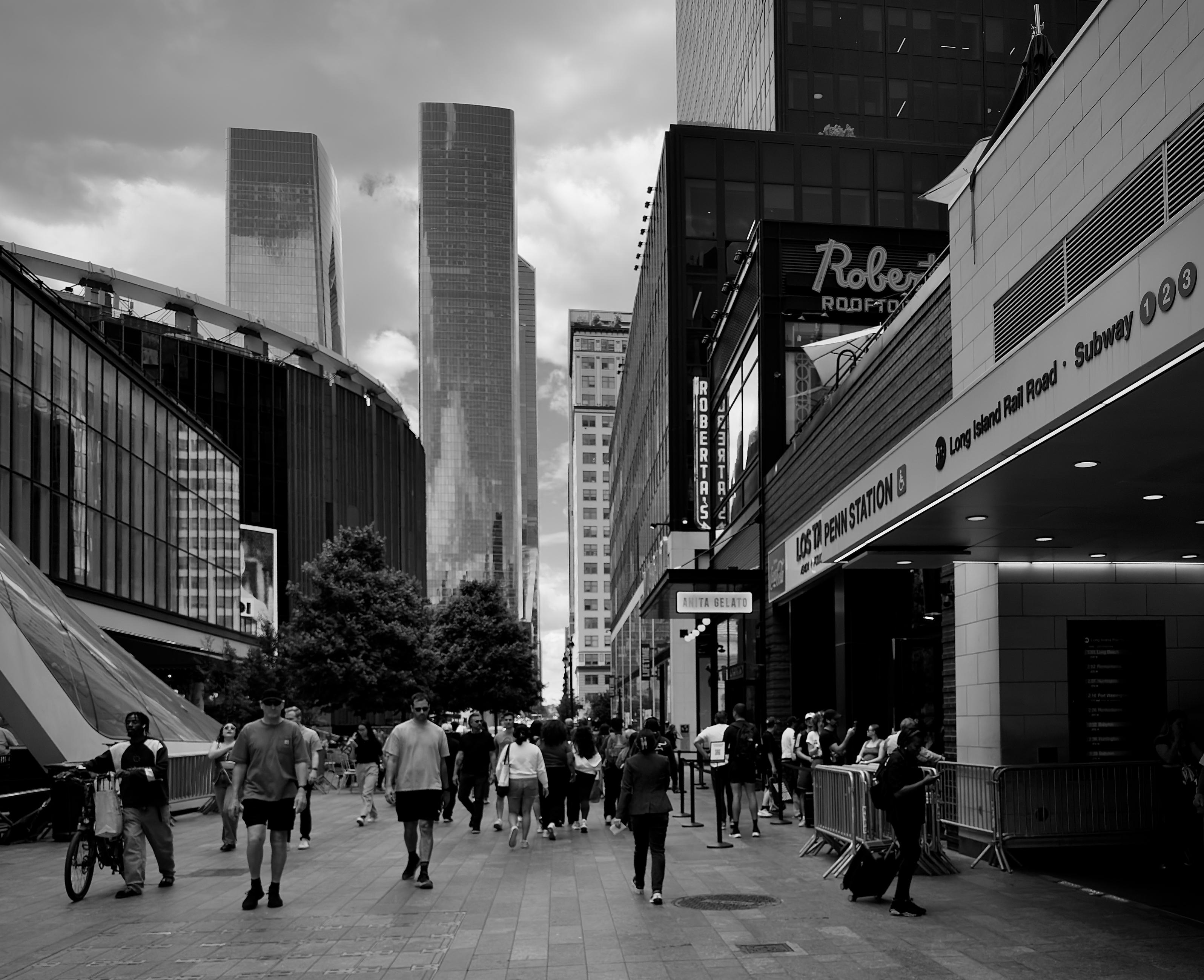 A noir, wide-angle photograph in square aspect ratio that captures the street scene outside of Penn Station in Manhattan. On the right, the entrance to Penn Station is prominent, featuring signage for the "Long Island Rail Road" and the "Subway 1, 2, 3" lines. The architecture is clean and contemporary, with a modern overhang and glass-paneled facades. Further down the street, a sign for "Roberta's" establishment is visible. In the background, striking, monolithic skyscrapers in Hudson Yards rise toward a cloudy, dramatic sky. A diverse group of people is captured in motion, some carrying bags. On the left side of the frame, the curved structure of Madison Square Garden looms, creating a sharp contrast between the older stadium design and the sleek, modern glass buildings nearby. The lighting and monochromatic treatment of the image heighten the textural details of the architecture.