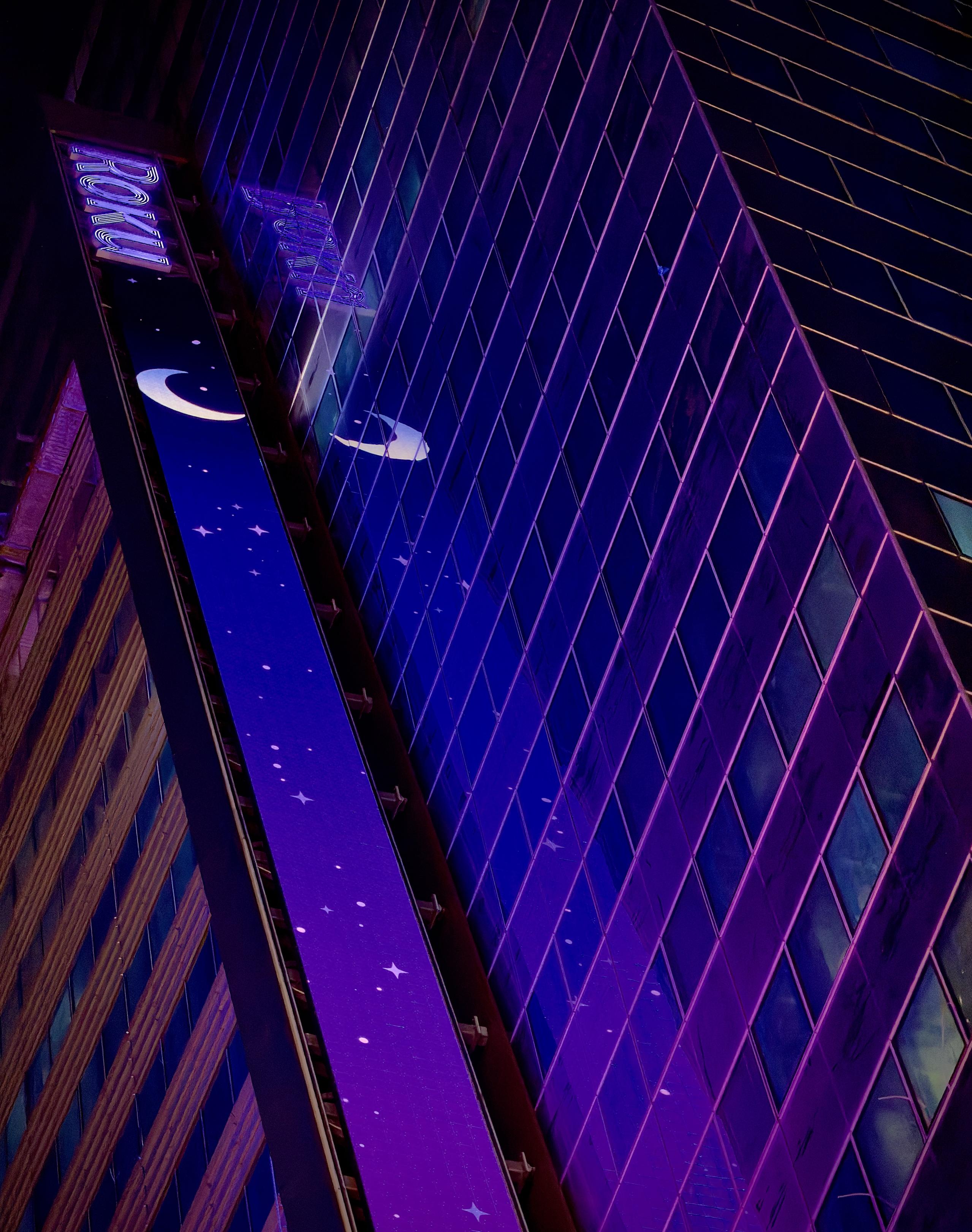A color, wide-angle photograph in portrait orientation taken at nighttime that captures a perspective of a modern skyscraper in Times Square, Manhattan, featuring prominent branding for Roku. A vertical, illuminated LED display runs along the side of the building, bathed in a vibrant shade of purple. The digital screen displays a whimsical night-sky theme, complete with a glowing crescent moon and various star icons scattered against the deep violet backdrop. In the upper portion of the frame, the illuminated "Roku" logo is clearly visible, anchored at the top of the vertical screen. The reflection of the crescent moon and the purple light can be seen faintly shimmering on the building's glass curtain wall.