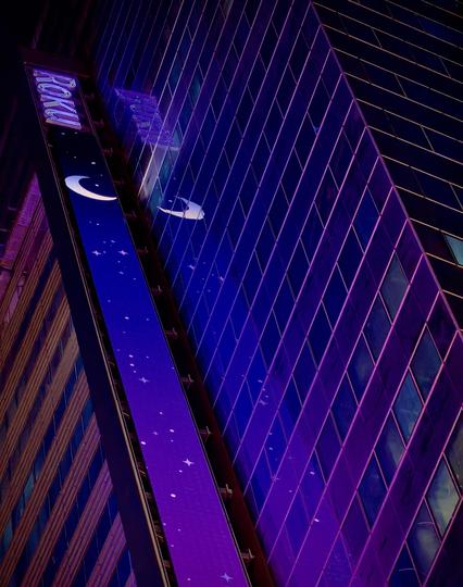 A color, wide-angle photograph in portrait orientation taken at nighttime that captures a perspective of a modern skyscraper in Times Square, Manhattan, featuring prominent branding for Roku. A vertical, illuminated LED display runs along the side of the building, bathed in a vibrant shade of purple. The digital screen displays a whimsical night-sky theme, complete with a glowing crescent moon and various star icons scattered against the deep violet backdrop. In the upper portion of the frame, the illuminated "Roku" logo is clearly visible, anchored at the top of the vertical screen. The reflection of the crescent moon and the purple light can be seen faintly shimmering on the building's glass curtain wall.