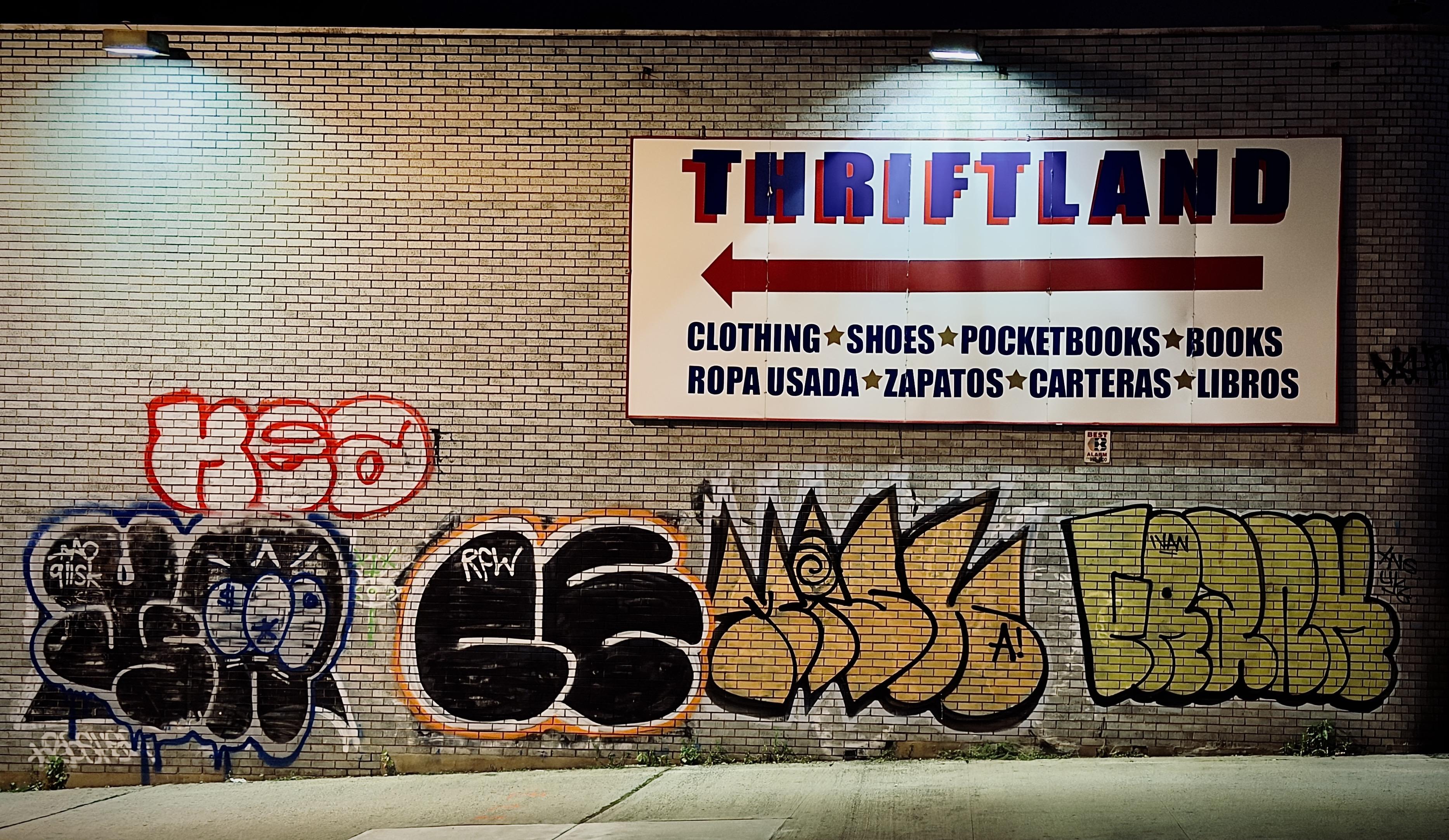 A color, wide-angle photograph in landscape orientation captured at night along River Avenue in the Bronx. A light-colored brick wall is covered with various forms of street art and signage. At the top right of the wall is a large, rectangular sign for "THRIFTLAND" with a red arrow pointing to the left, below which it lists "CLOTHING SHOES POCKETBOOKS BOOKS" and their Spanish equivalents in dark blue text. The brick surface beneath the sign is adorned with several distinct graffiti tags. One notable tag in red outline sits above another with blue accents, while a series of other stylized, overlapping graffiti designs in black, yellow, and green span the lower section of the wall. Two small, cool-toned light fixtures are mounted near the top edge of the brick wall, casting downward illumination that creates subtle highlights on the textured brickwork. A narrow concrete sidewalk runs along the base of the wall, growing in height from left to right as the incline of the hill steepens.