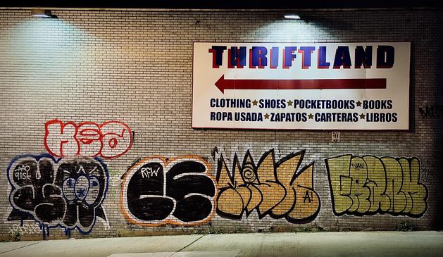 A color, wide-angle photograph in landscape orientation captured at night along River Avenue in the Bronx. A light-colored brick wall is covered with various forms of street art and signage. At the top right of the wall is a large, rectangular sign for "THRIFTLAND" with a red arrow pointing to the left, below which it lists "CLOTHING SHOES POCKETBOOKS BOOKS" and their Spanish equivalents in dark blue text. The brick surface beneath the sign is adorned with several distinct graffiti tags. One notable tag in red outline sits above another with blue accents, while a series of other stylized, overlapping graffiti designs in black, yellow, and green span the lower section of the wall. Two small, cool-toned light fixtures are mounted near the top edge of the brick wall, casting downward illumination that creates subtle highlights on the textured brickwork. A narrow concrete sidewalk runs along the base of the wall, growing in height from left to right as the incline of the hill steepens.
