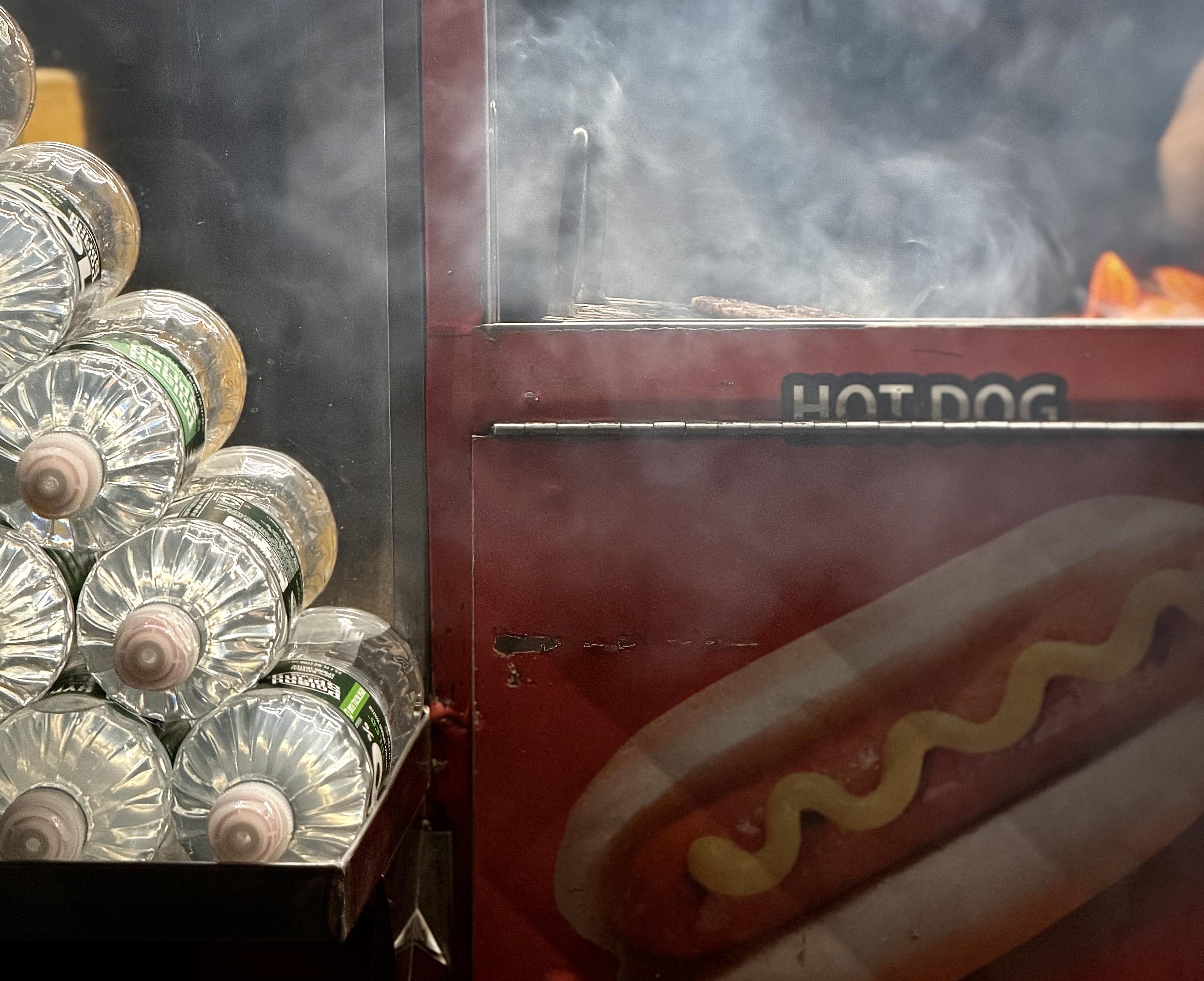 A color, medium-angle photograph in landscape orientation that captures the street scene outside of Grand Central Terminal at night. The focal point of the image is the side of a red hot dog food cart. On the left side of the frame, a pyramid shaped stack of clear plastic water bottles is arranged neatly in a metal holder lying on their sides, with the ribbed textures of the bottles and their caps prominently displayed. The right side of the image features the cart's exterior, which is painted a deep red and displays a graphic of a hot dog topped with a wavy line of yellow mustard. Above the graphic, the words "HOT DOG" are printed in a stylized, bold font. A partition separates the viewer from the cooking area, where thick, swirling white steam rises from the grill. The blurred shapes of hot dogs and sausages are visible cooking on the metal grate, surrounded by the haze of the steam. The overall lighting is m dim from overhead spotlights in the nighttime atmosphere.
