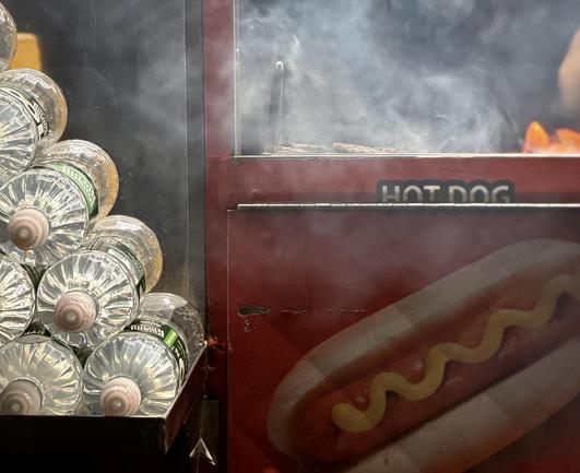 A color, medium-angle photograph in landscape orientation that captures the street scene outside of Grand Central Terminal at night. The focal point of the image is the side of a red hot dog food cart. On the left side of the frame, a pyramid shaped stack of clear plastic water bottles is arranged neatly in a metal holder lying on their sides, with the ribbed textures of the bottles and their caps prominently displayed. The right side of the image features the cart's exterior, which is painted a deep red and displays a graphic of a hot dog topped with a wavy line of yellow mustard. Above the graphic, the words "HOT DOG" are printed in a stylized, bold font. A partition separates the viewer from the cooking area, where thick, swirling white steam rises from the grill. The blurred shapes of hot dogs and sausages are visible cooking on the metal grate, surrounded by the haze of the steam. The overall lighting is m dim from overhead spotlights in the nighttime atmosphere.