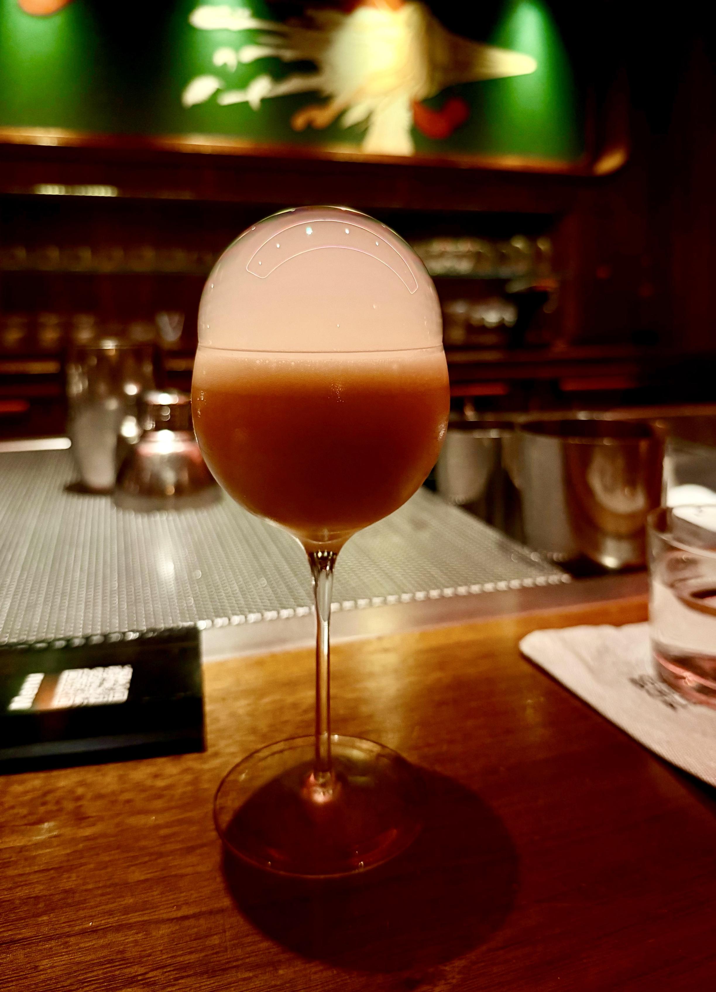 A color, medium-angle photograph in portrait orientation that captures a beautifully crafted cocktail known as the "Custard Bun" from Double Chicken Please cocktail bar on the lower east side of Manhattan. The drink is presented in an elegant, long-stemmed wine glass centered on a dark wooden bar top. The drink itself is an opaque, warm amber-tan color, topped with a prominent, translucent bubble dome that rests on the rim of the glass. The lighting is intimate and moody, typical of a high-end cocktail bar setting. The background is softly blurred, showing the warm, dimly lit interior of the bar. A dark green panel with faint, abstract decorative artwork is visible behind the bar, along with glimpses of stainless-steel bar equipment and glassware on a textured mat. To the right, a folded napkin and a small glass of water sit on the bar, while a menu rests to the left.